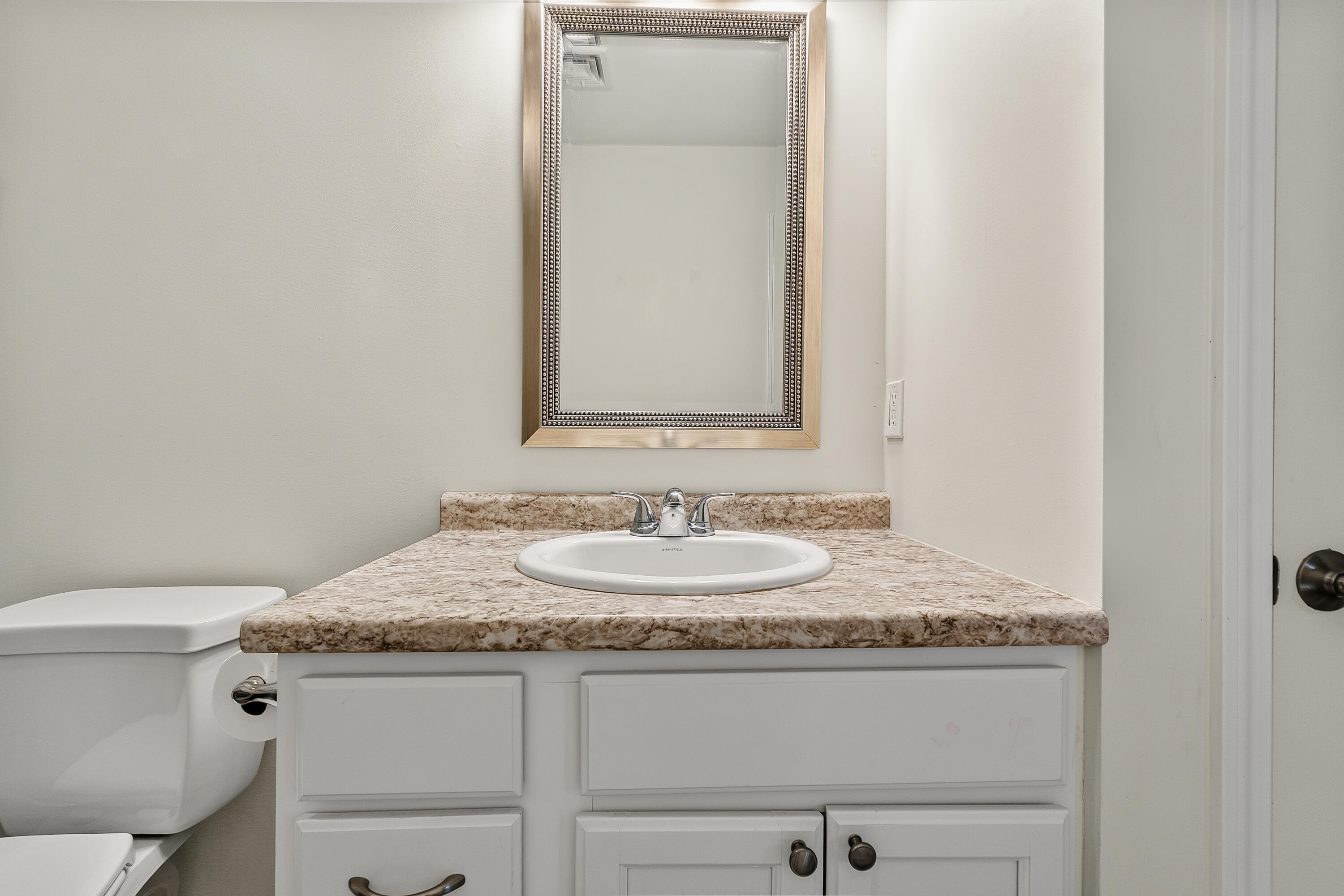 Bathroom vanity with an oval sink, mottled beige countertop, framed mirror, and adjacent toilet.