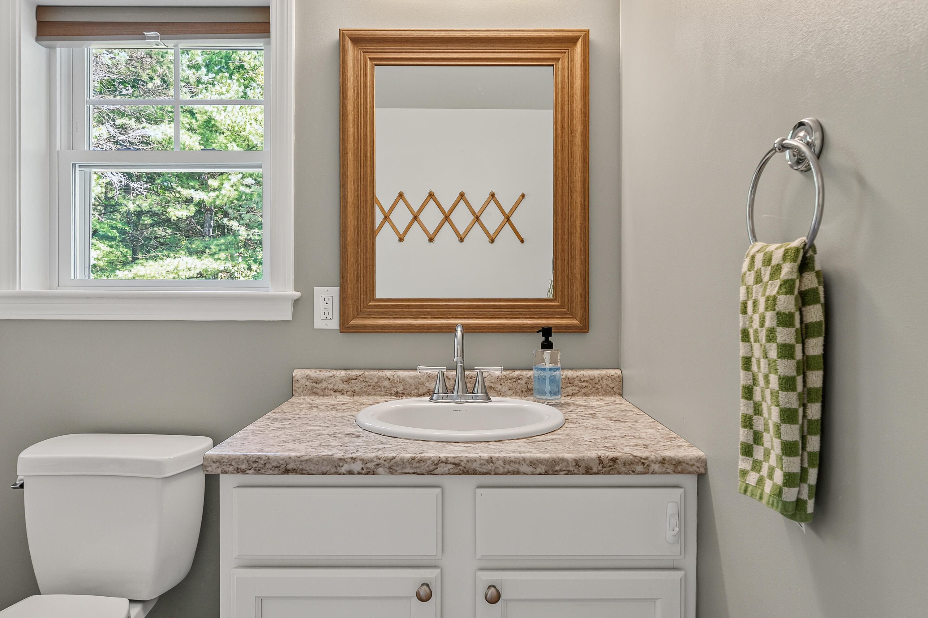 Bathroom vanity with a wood-framed mirror, window overlooking trees, and green checkered towel.