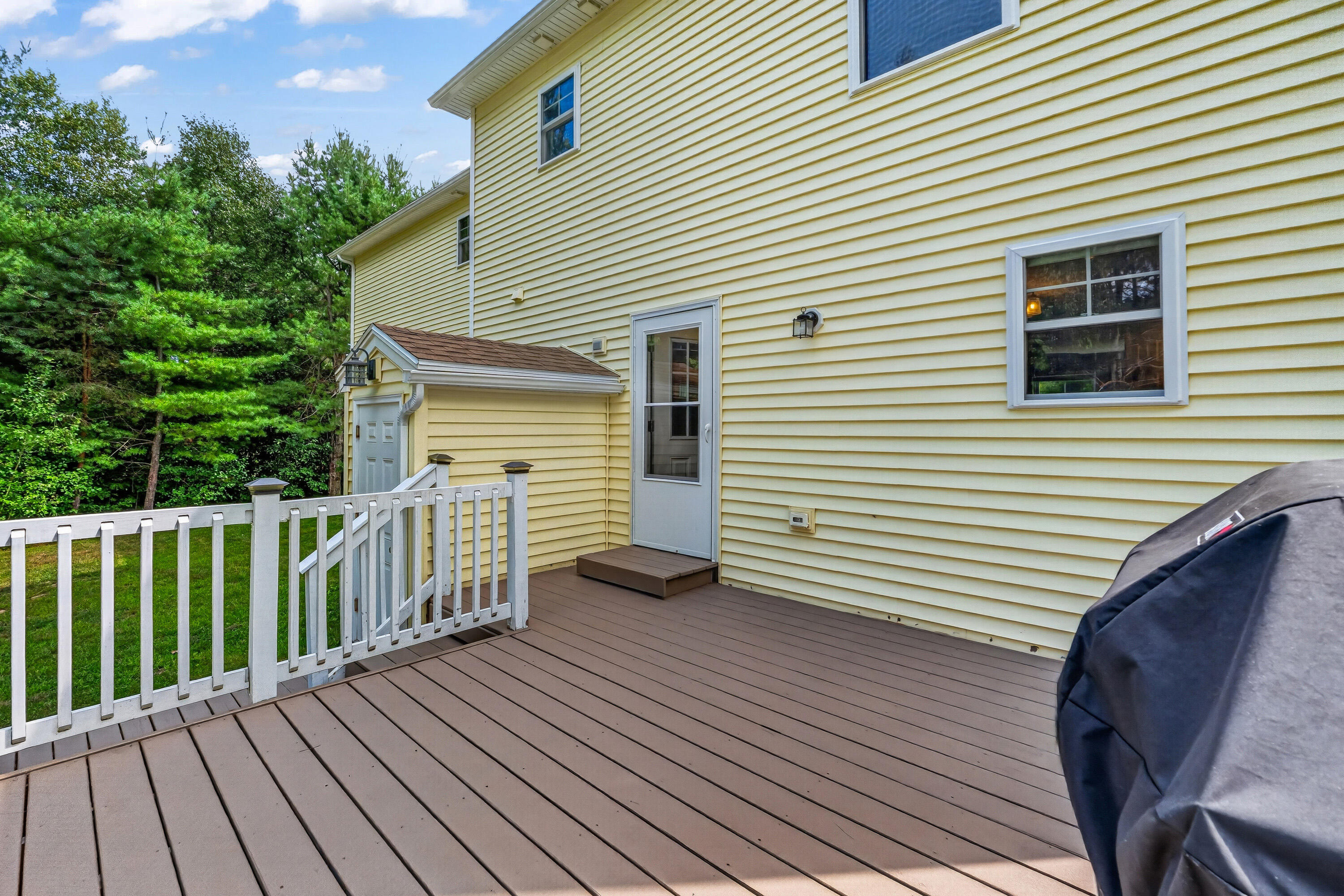 Brown deck with white railing beside a yellow-sided house, overlooking a wooded yard.