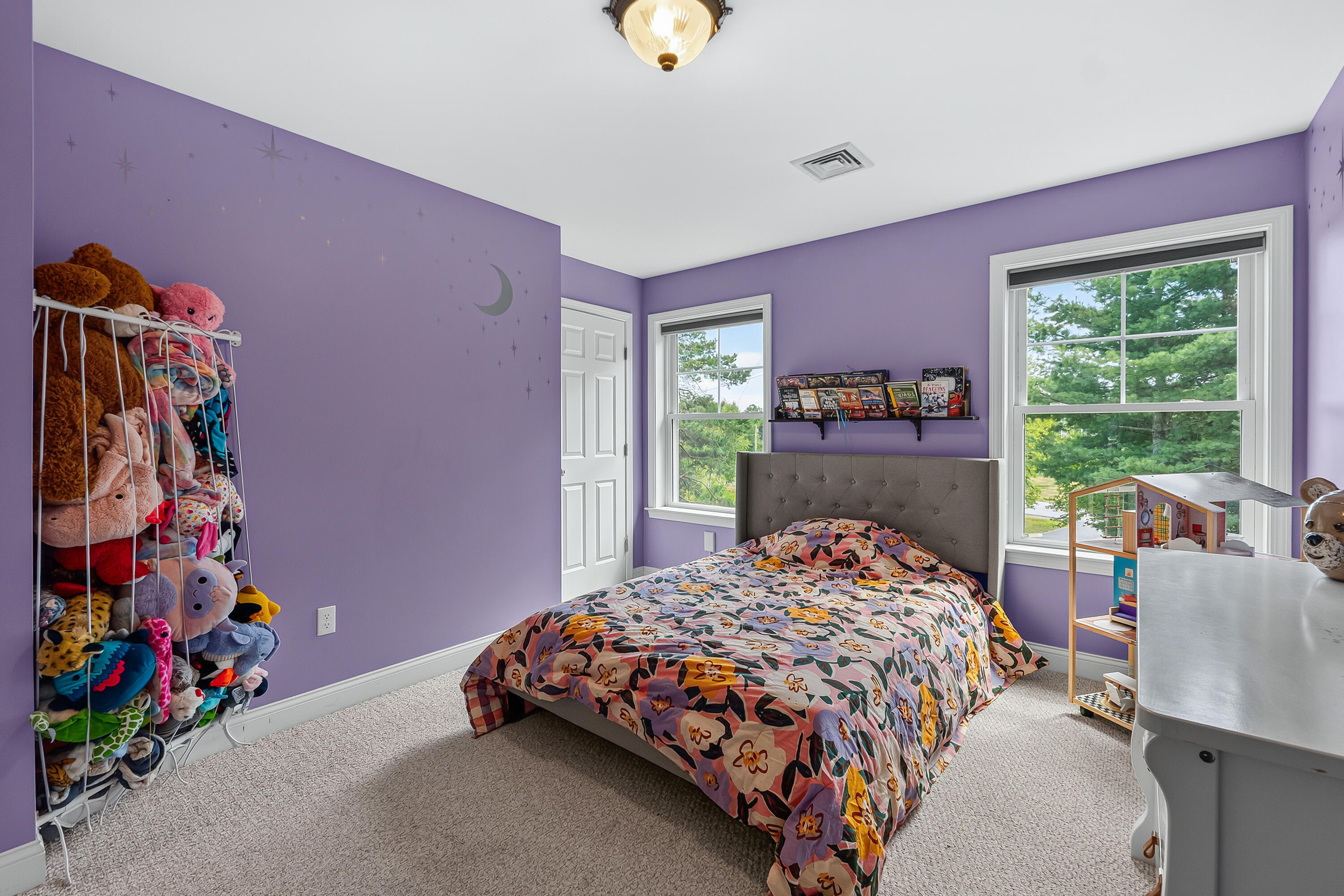 Lavender bedroom with a floral-covered bed, stuffed animals, and two windows overlooking trees.