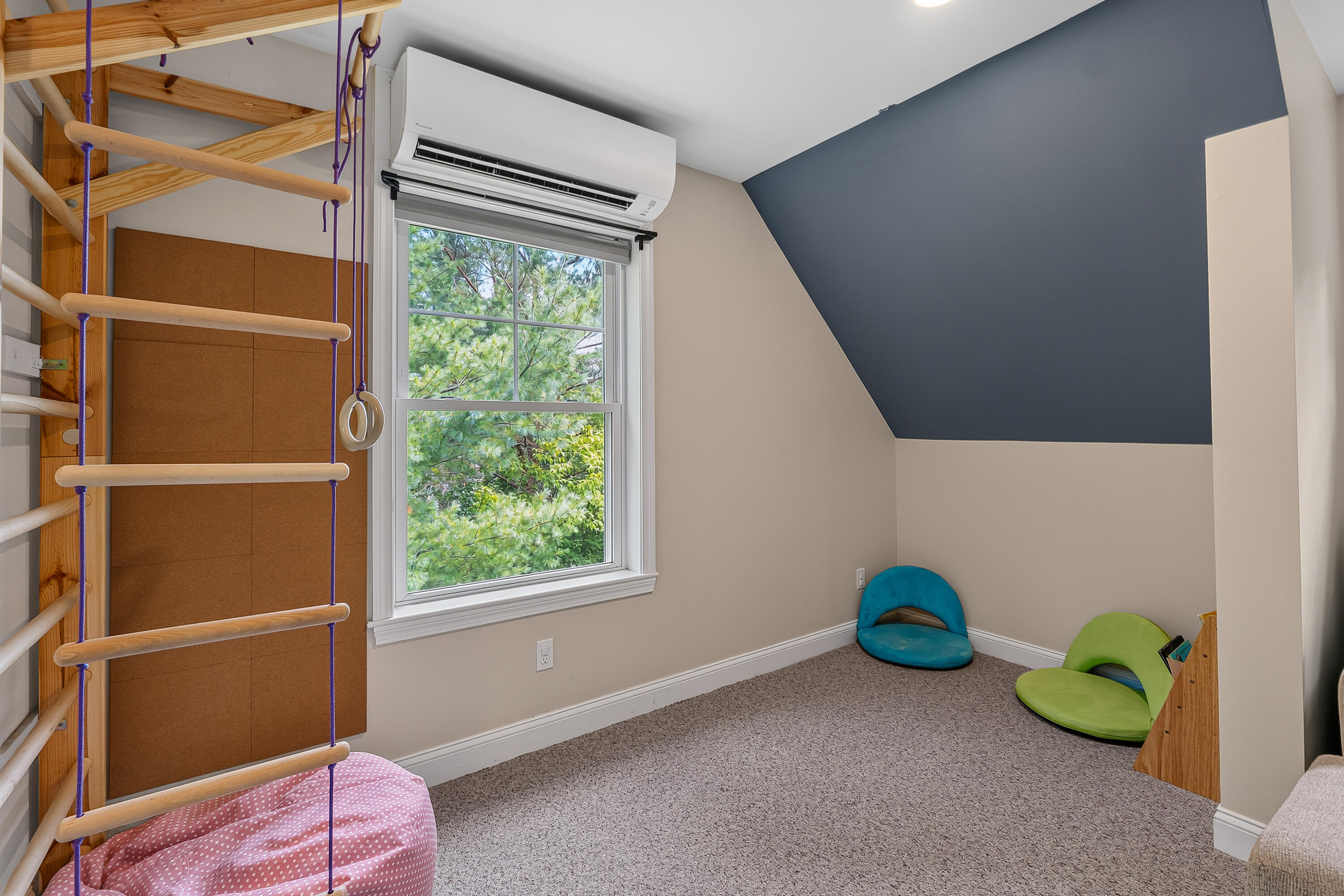 Attic playroom with wooden climbing bars, colorful floor chairs, and a window overlooking trees.