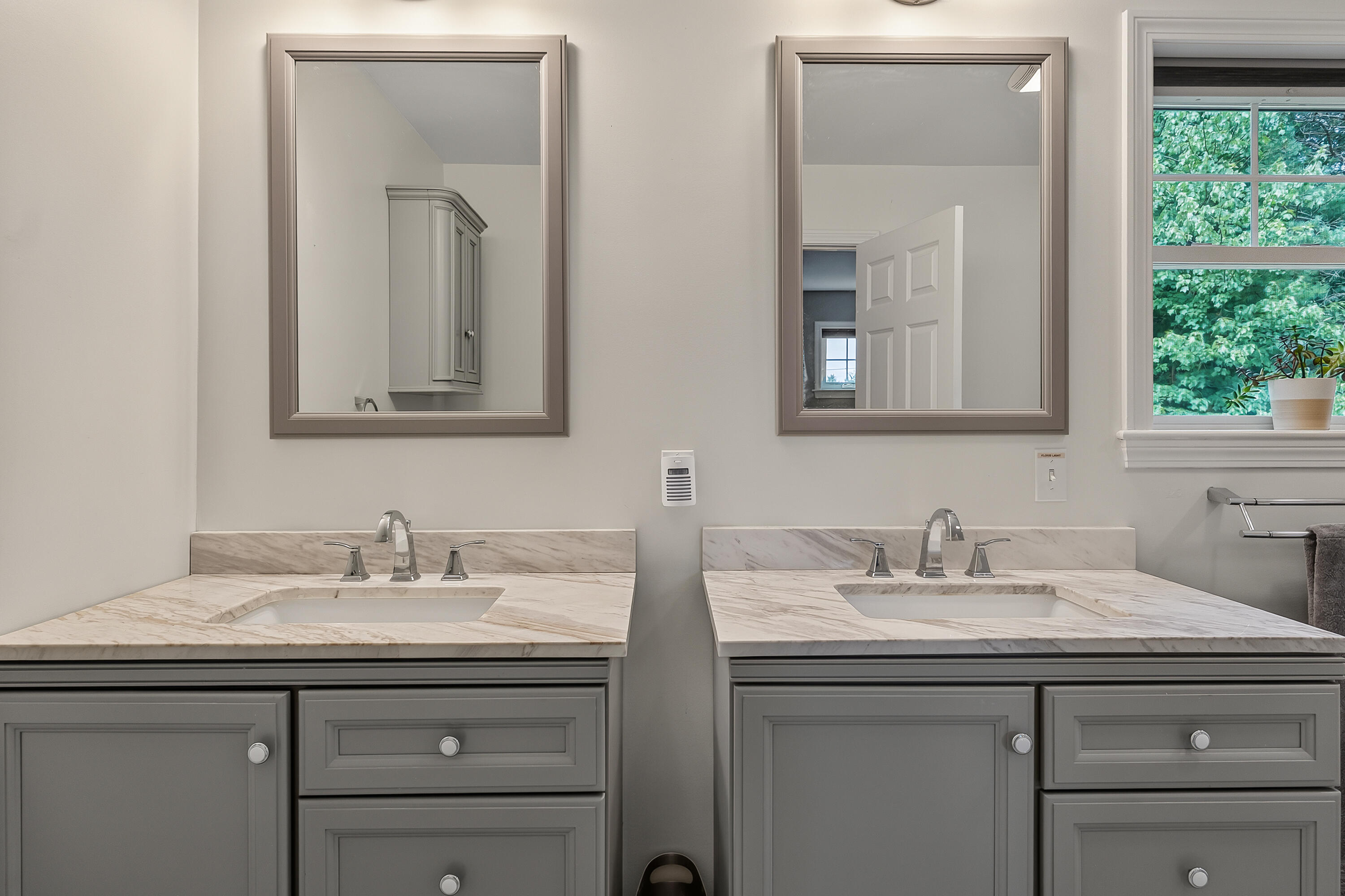 Bathroom double vanity with marble counters, gray cabinetry, framed mirrors, and a window overlooking greenery.