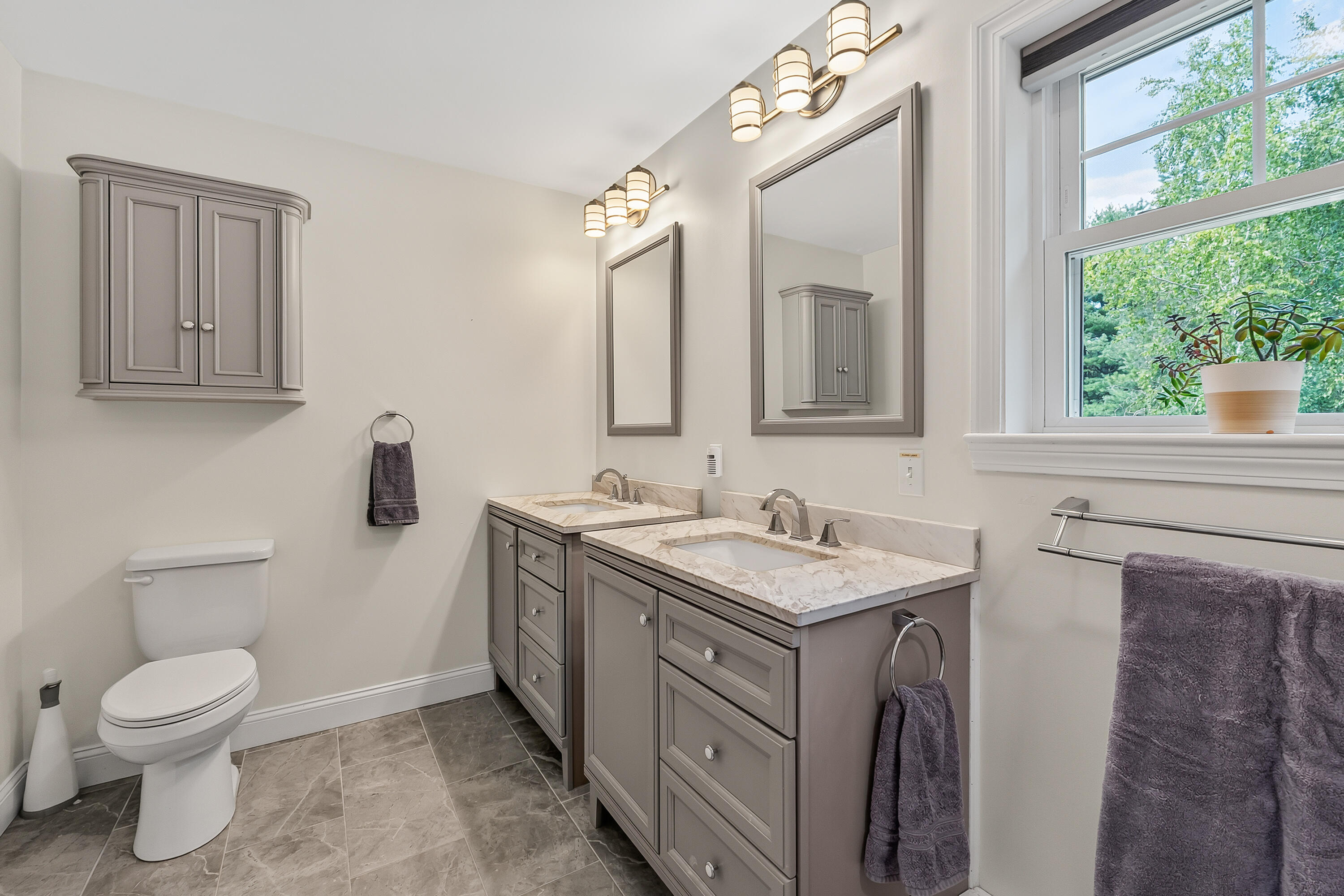 Bathroom with a gray double vanity, marble-look counters, and a large window overlooking trees.