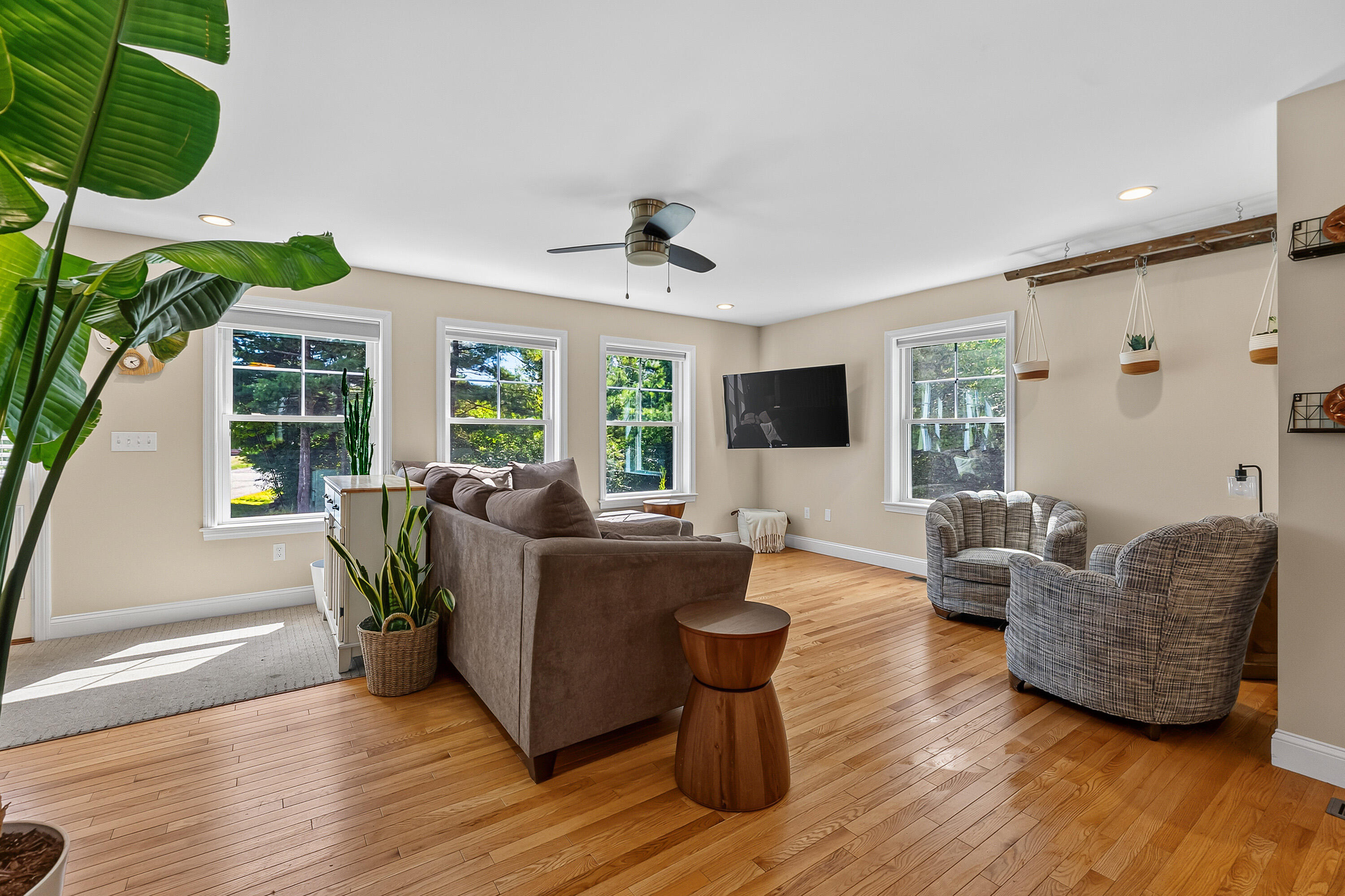 Living room with hardwood floors, a gray sofa, patterned armchairs, and large windows overlooking trees.