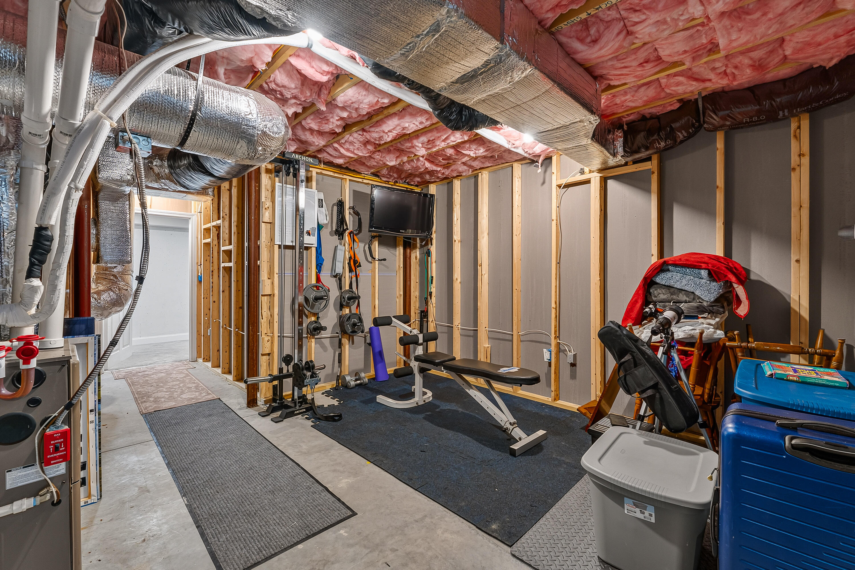 Basement gym with weight equipment and bench beneath exposed framing and insulation.