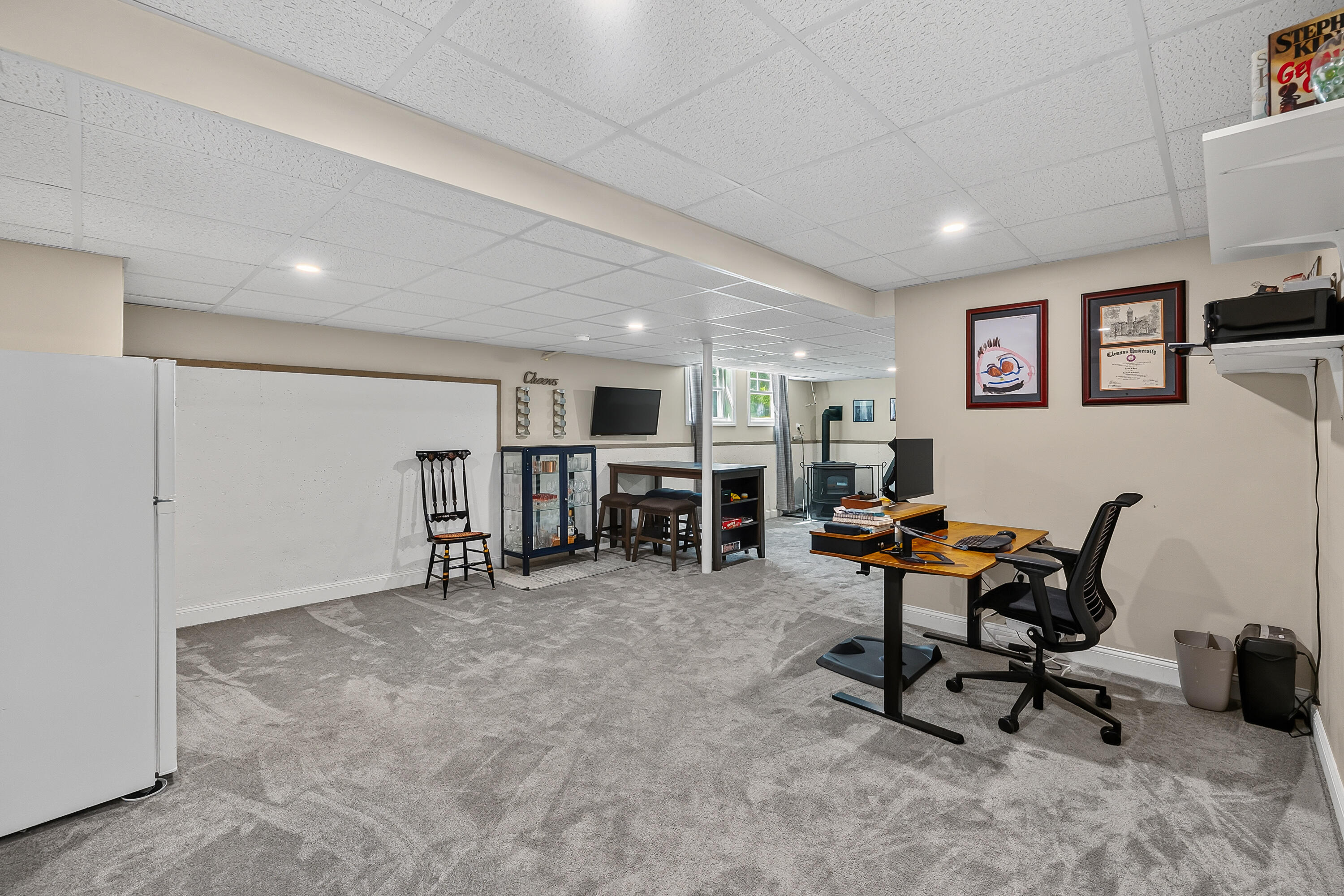 Furnished basement office with a desk, gray carpet, refrigerator, and television.