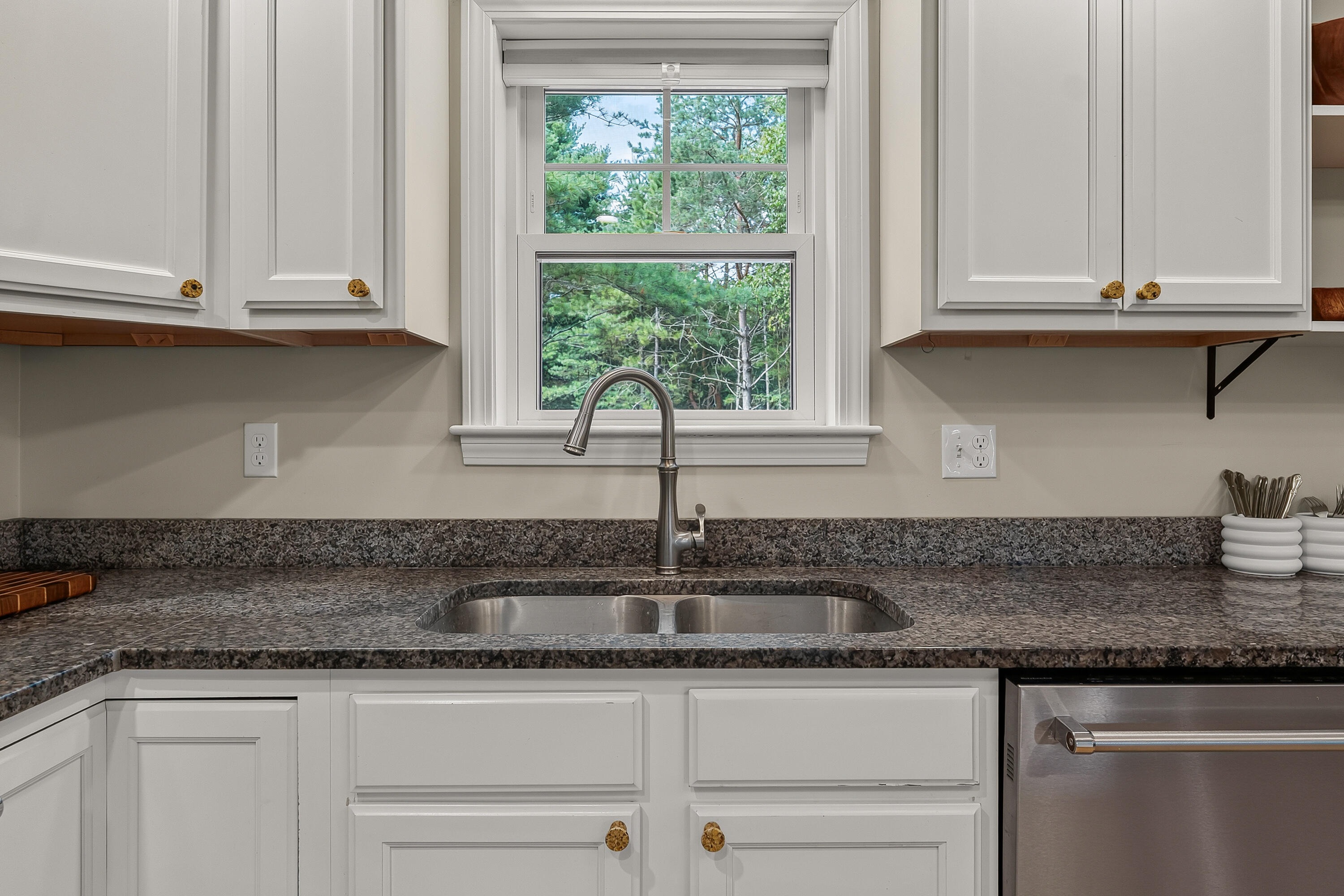 Kitchen sink beneath a window overlooking trees, framed by white cabinets and dark granite counters.