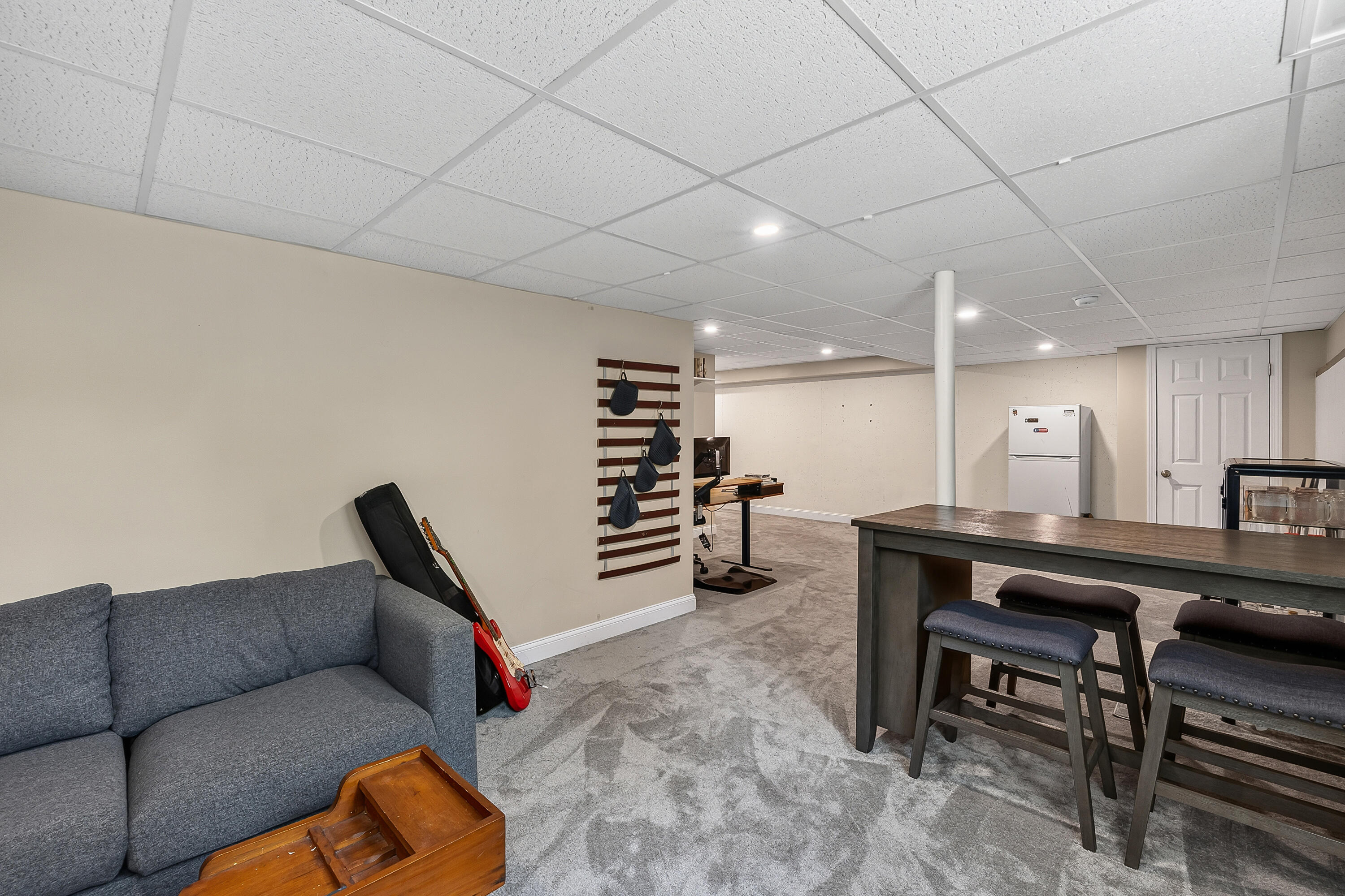 Furnished basement with a gray sectional, guitar, desk, mini fridge, and counter-height table with stools.