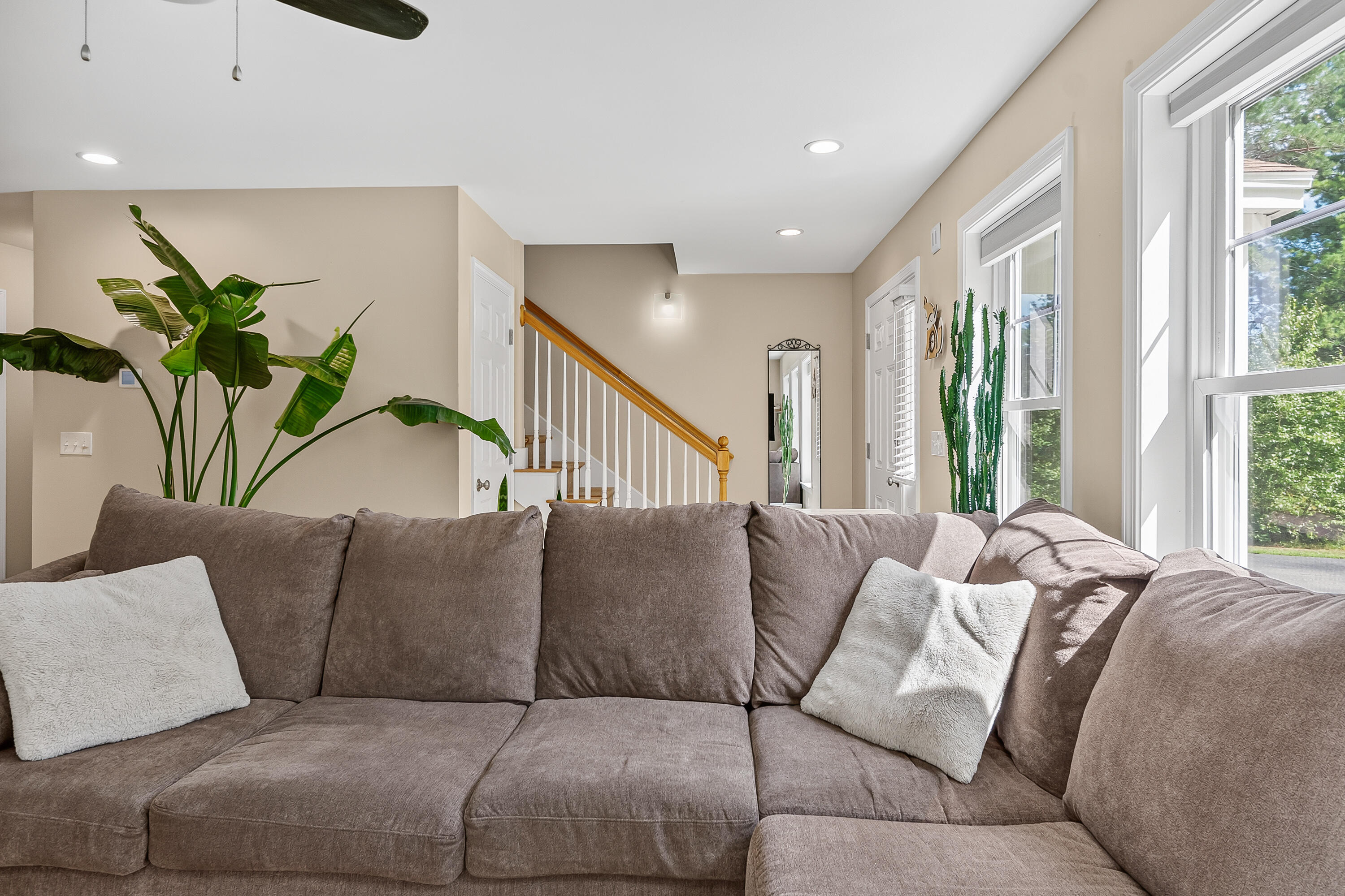 Furnished living room with a taupe sectional, tall windows, indoor plants, and a staircase.