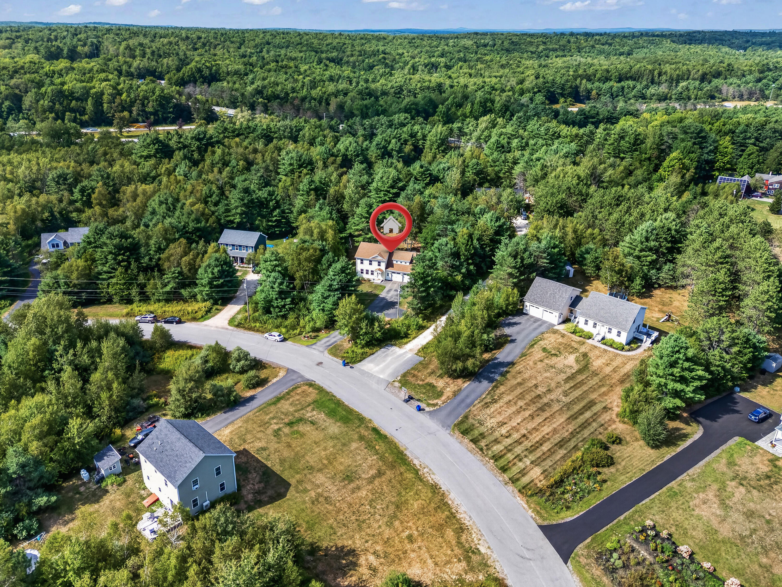 Aerial view of a white house marked by a red pin amid dense woods and nearby homes.