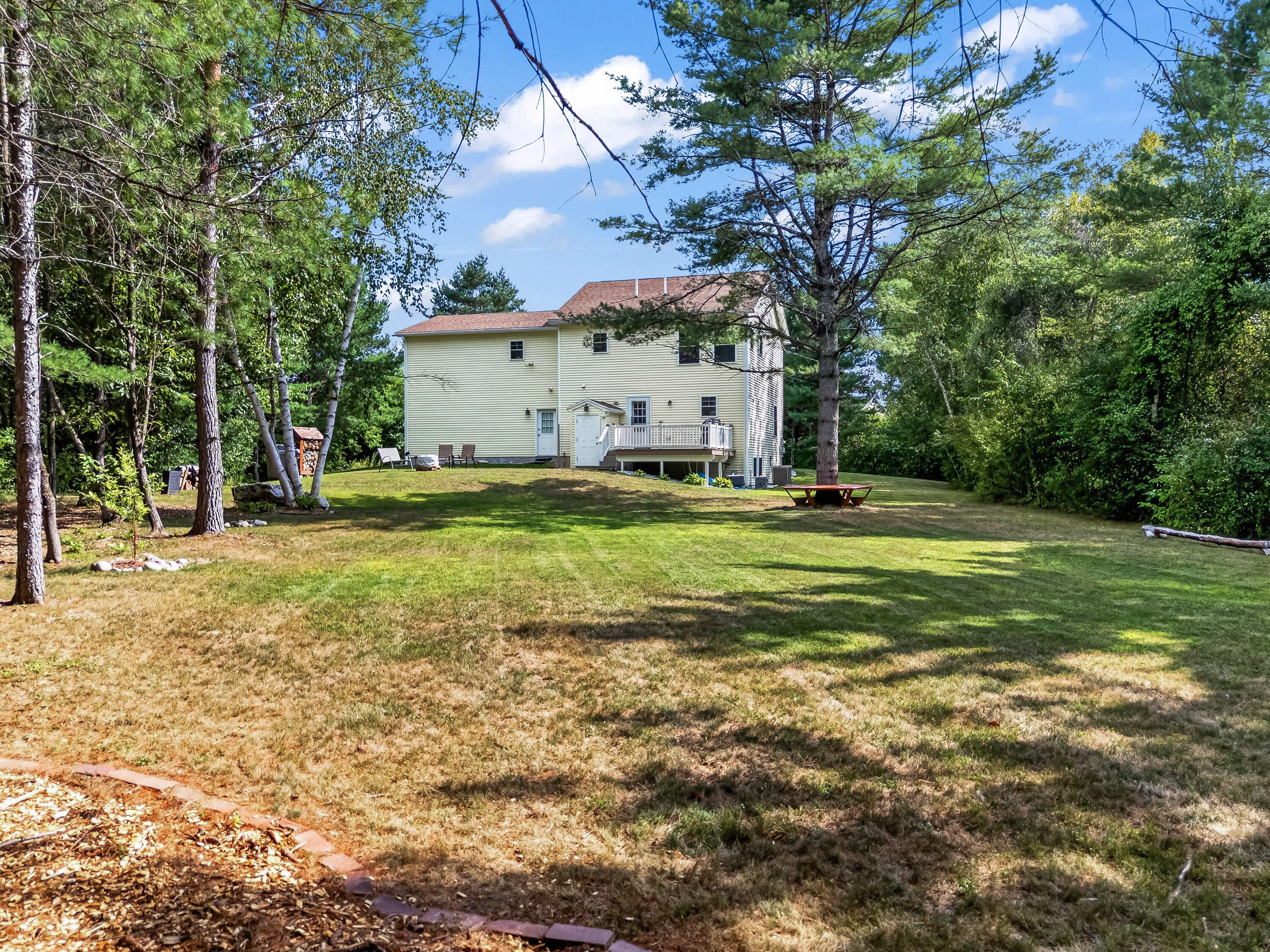 Pale yellow two-story house and raised deck beyond a broad lawn shaded by mature trees.