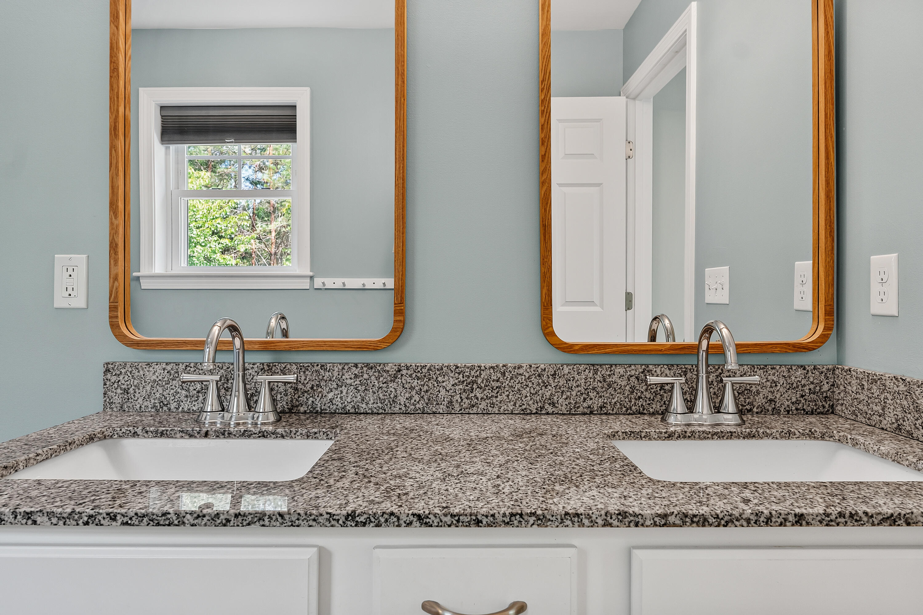 Bathroom double vanity with granite counters, two wood-framed mirrors, and a window reflected in one mirror.