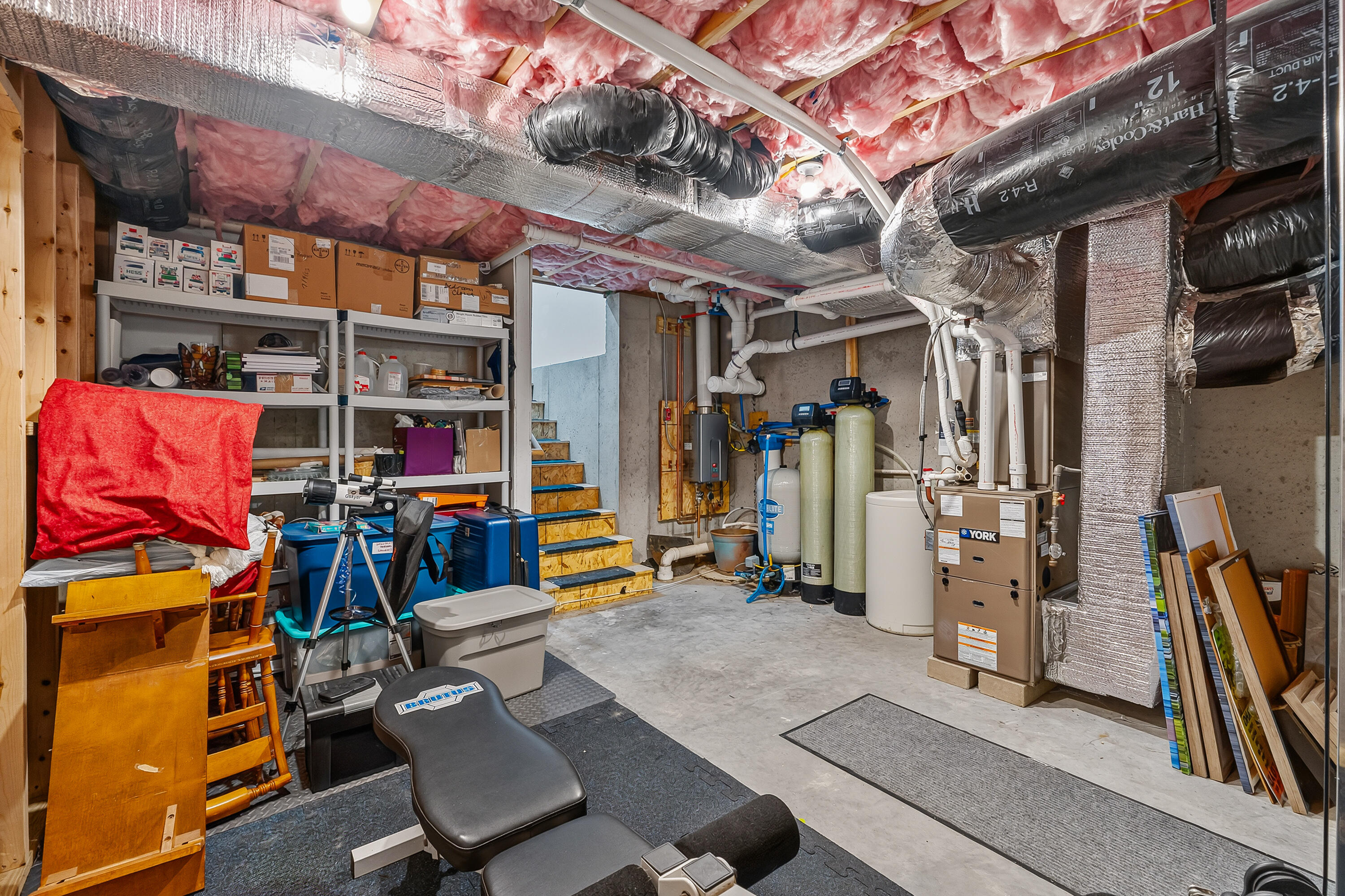 Basement utility room with exposed insulation, extensive ductwork, water treatment tanks, and stored exercise equipment.