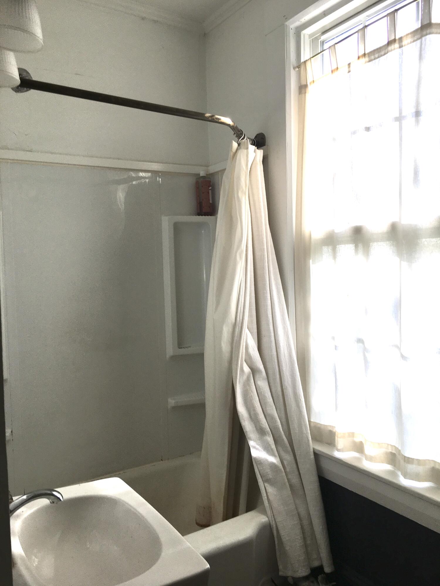 Bathroom tub and shower with a white curtain beside a bright window.
