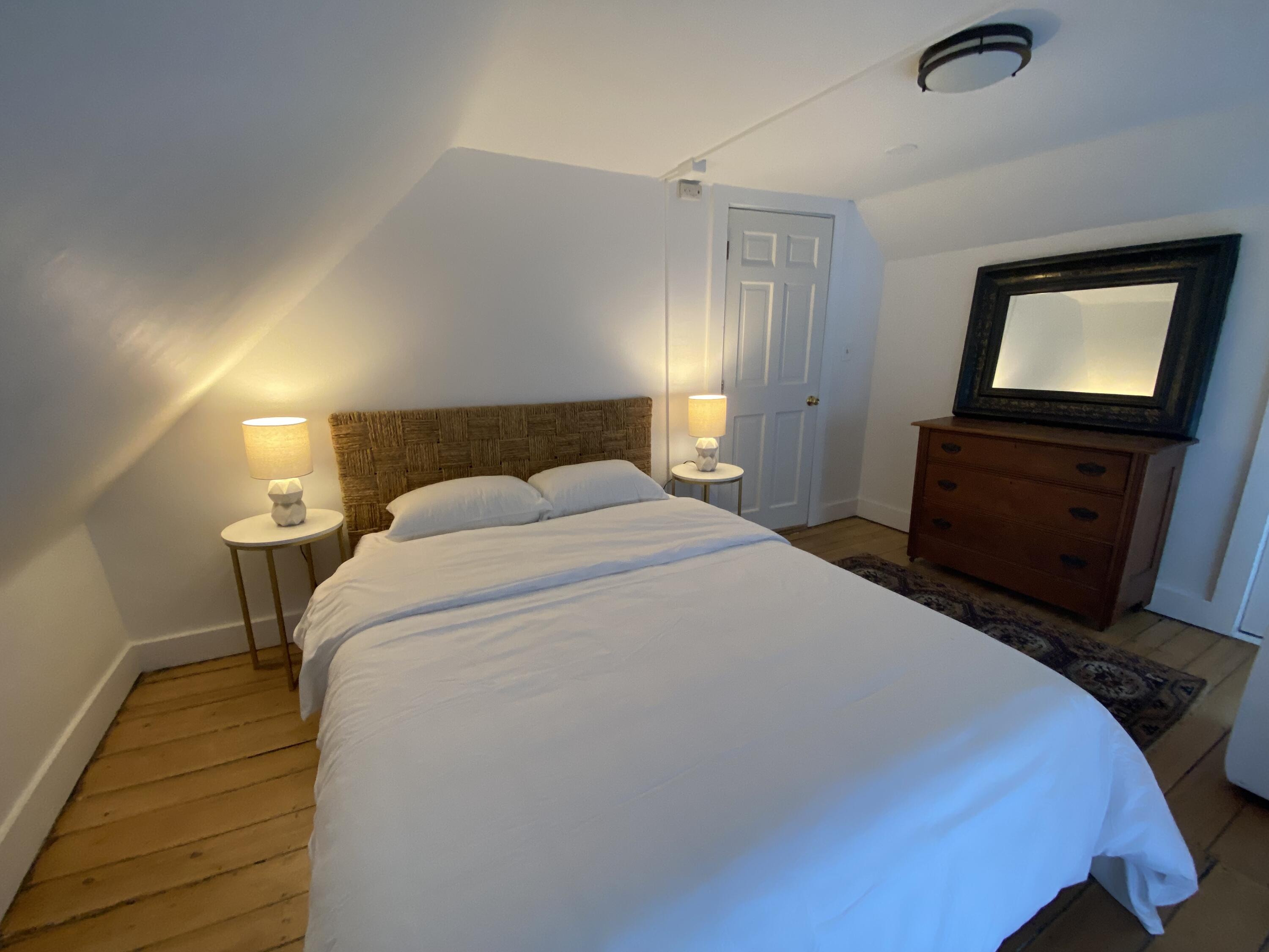 Bedroom with a white bed, woven headboard, wood floors, and a dresser topped by a large mirror.