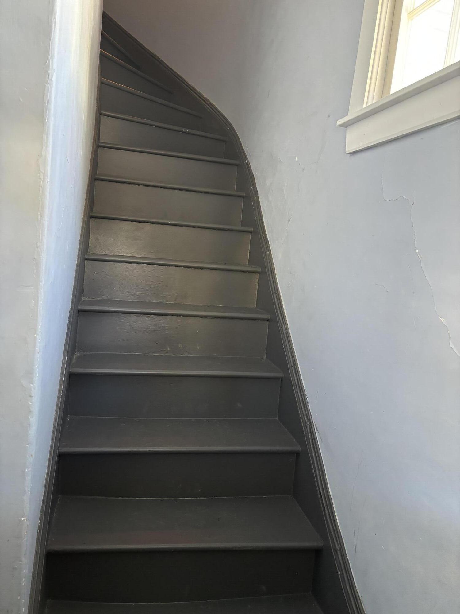 Narrow dark staircase curves upward beside a daylight window; cracks mark the pale blue-gray wall.