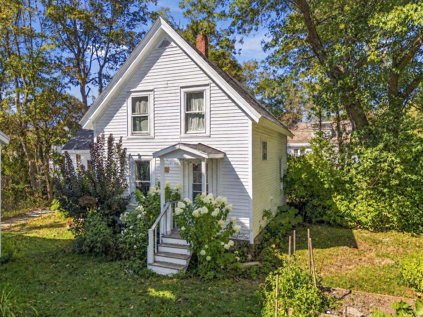 White clapboard house with a steep gable, covered entry, and lush garden surroundings.