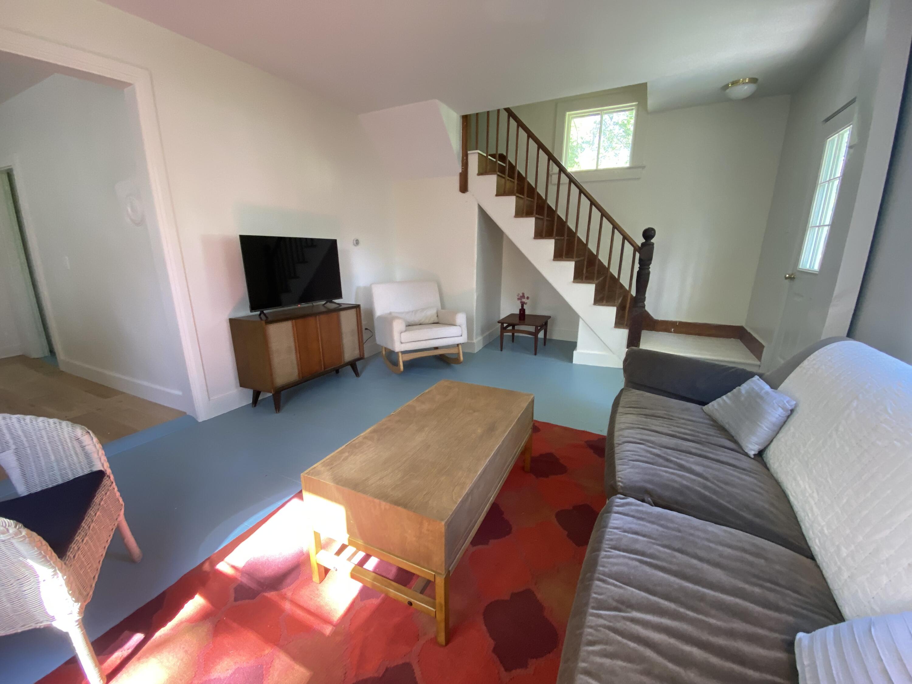 Living room with a blue floor, red patterned rug, gray sofa, and wooden staircase.