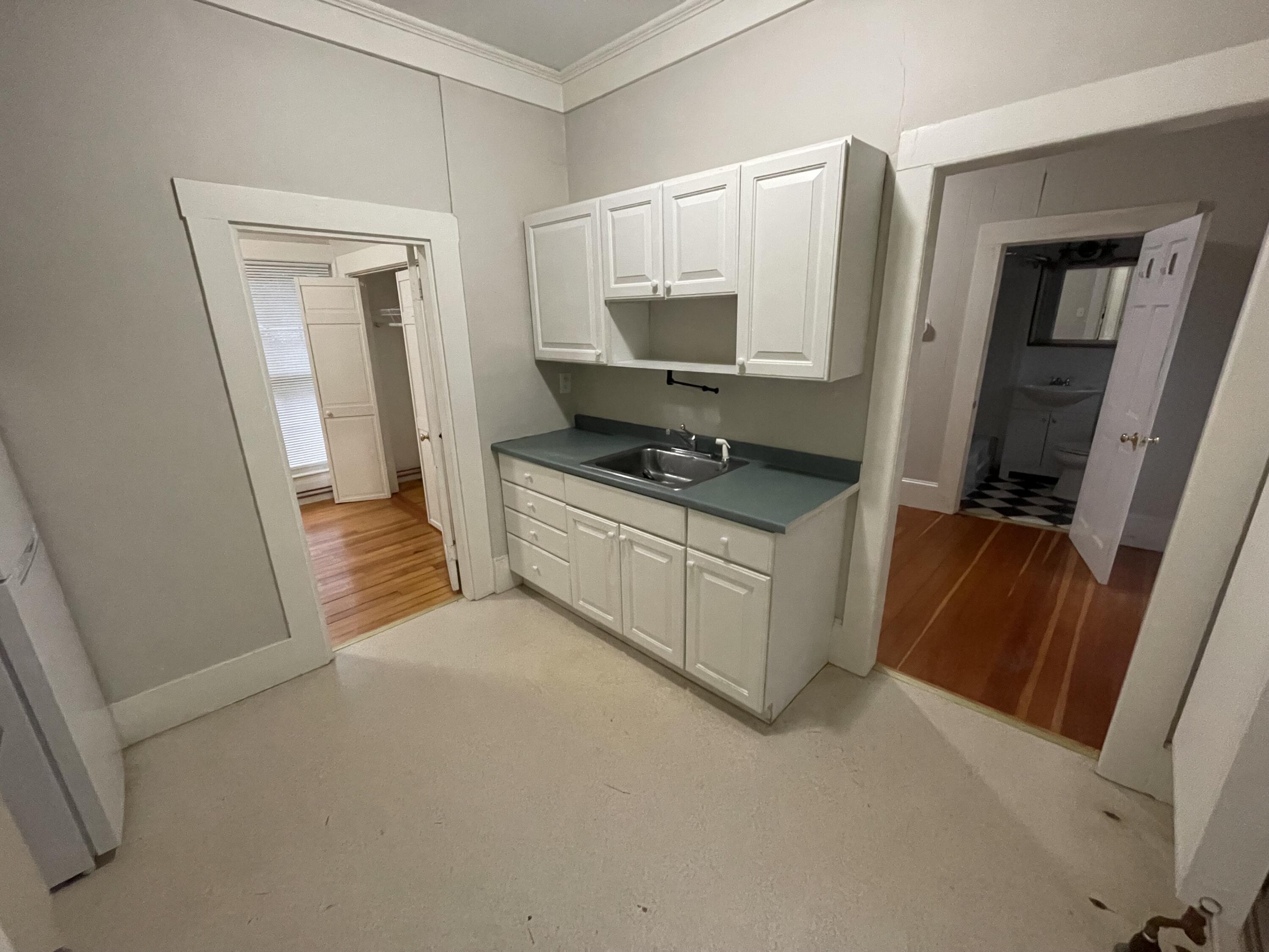 Vacant kitchen with white cabinets, gray countertop, and stainless-steel sink beside a bathroom doorway.