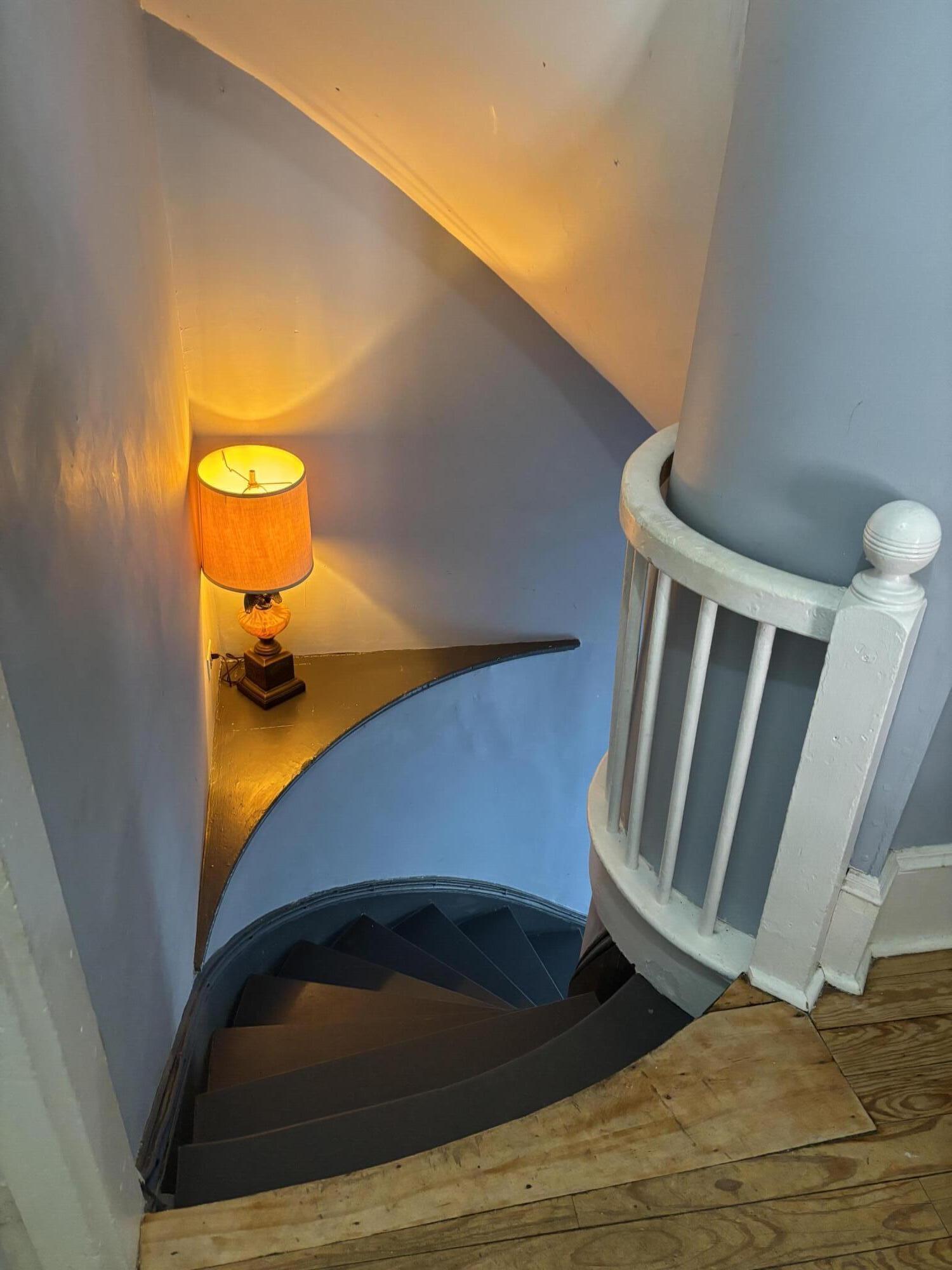 Curved dark staircase with a white balustrade and a glowing lamp on a narrow shelf.