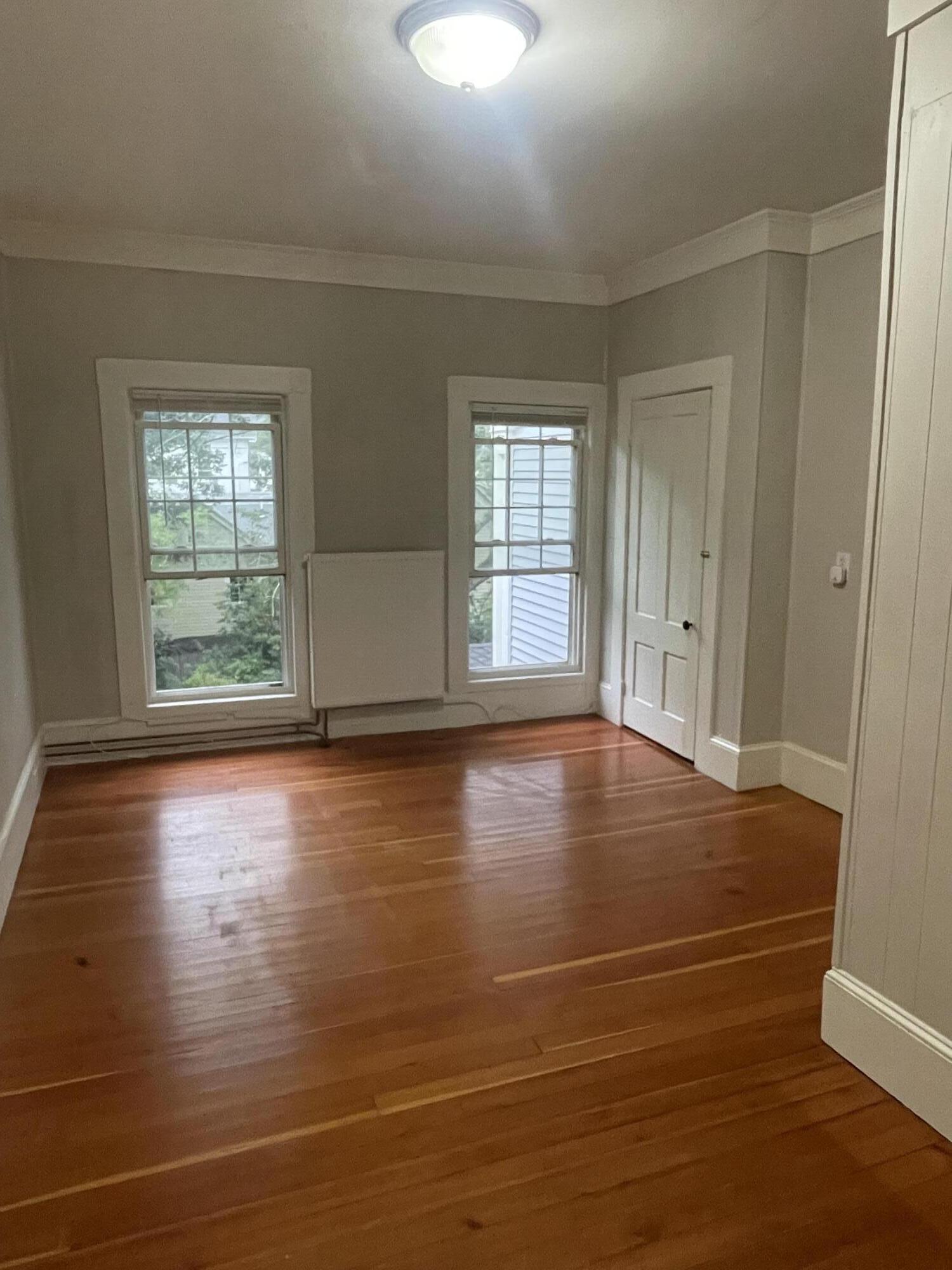 Empty room with polished hardwood floors, two windows, and a radiator beneath white trim.