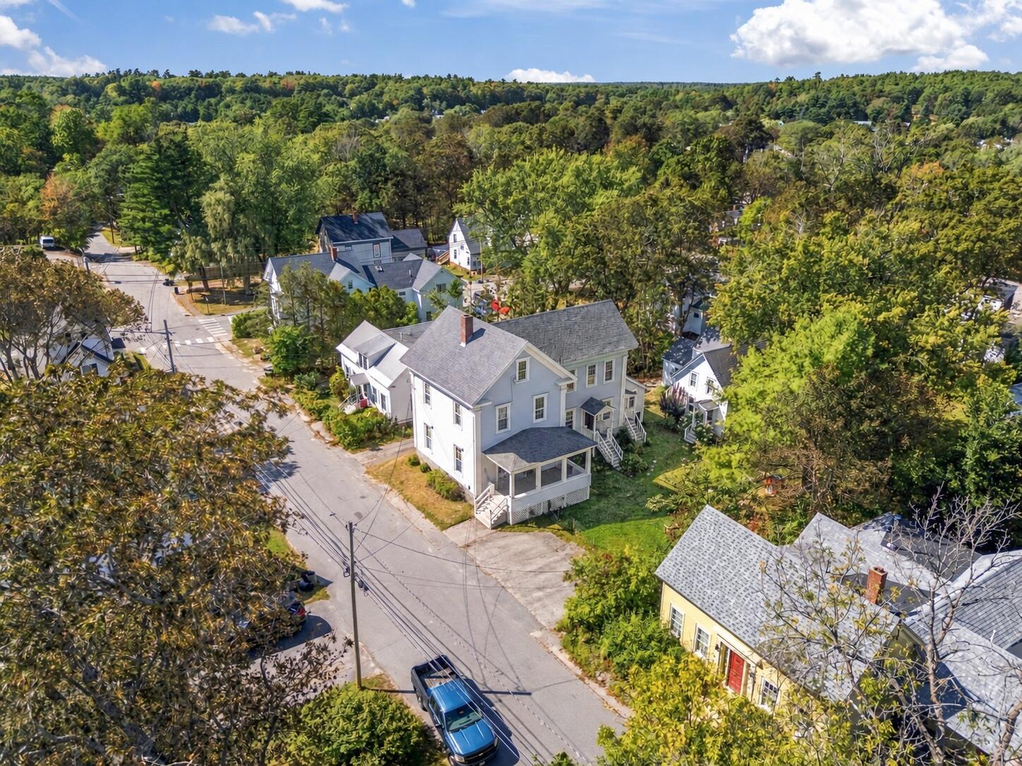 Aerial view of a light-colored multi-story house at a leafy street intersection, surrounded by neighboring homes.