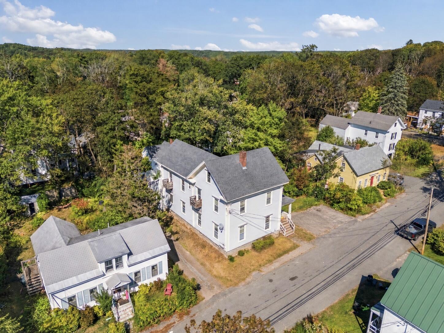 Aerial view of a white multi-story house beside a residential street, with wooded hills beyond the neighborhood.