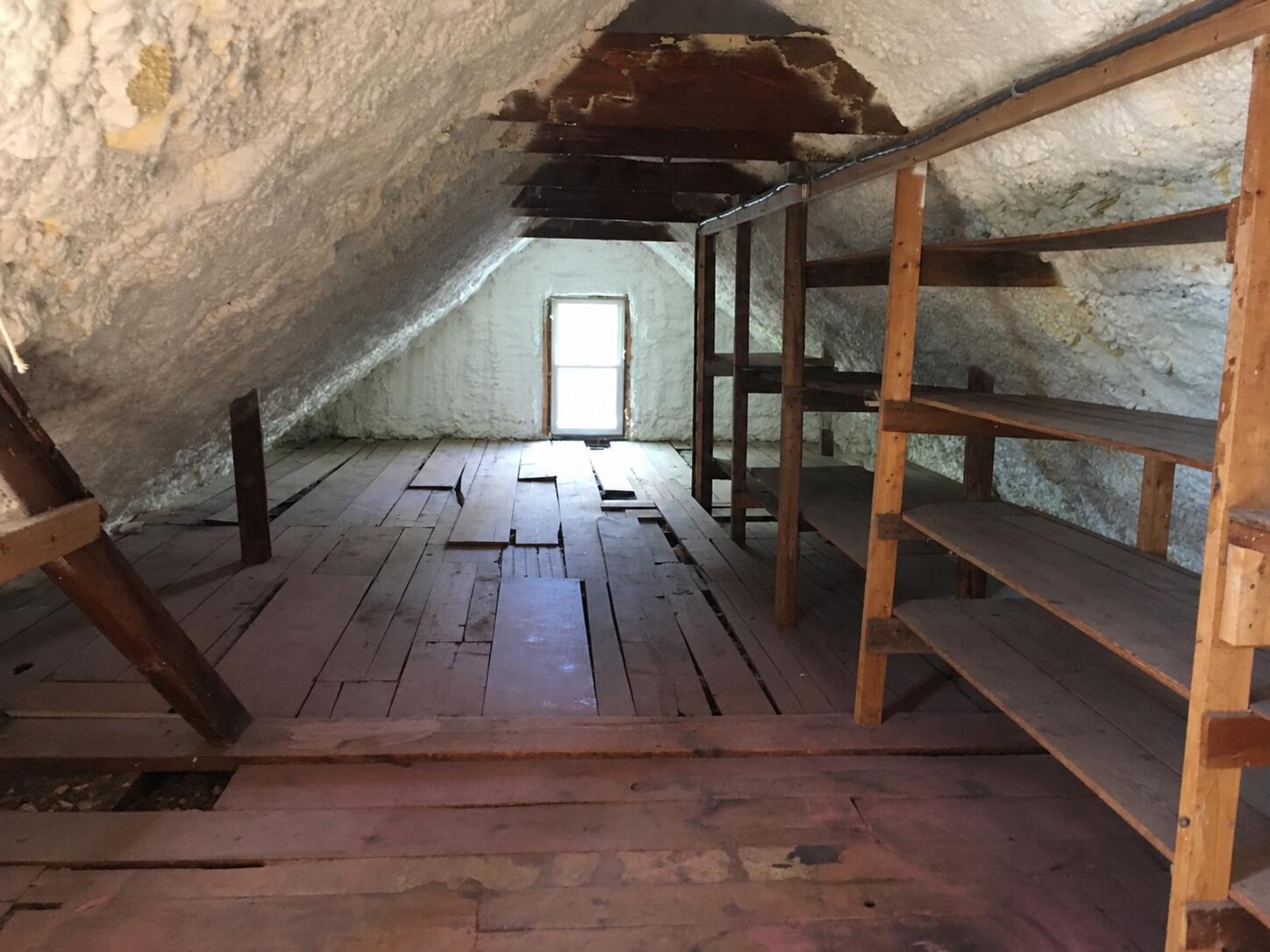 Unfinished attic with spray foam insulation, exposed framing, rough plank floors, and wooden storage shelves.