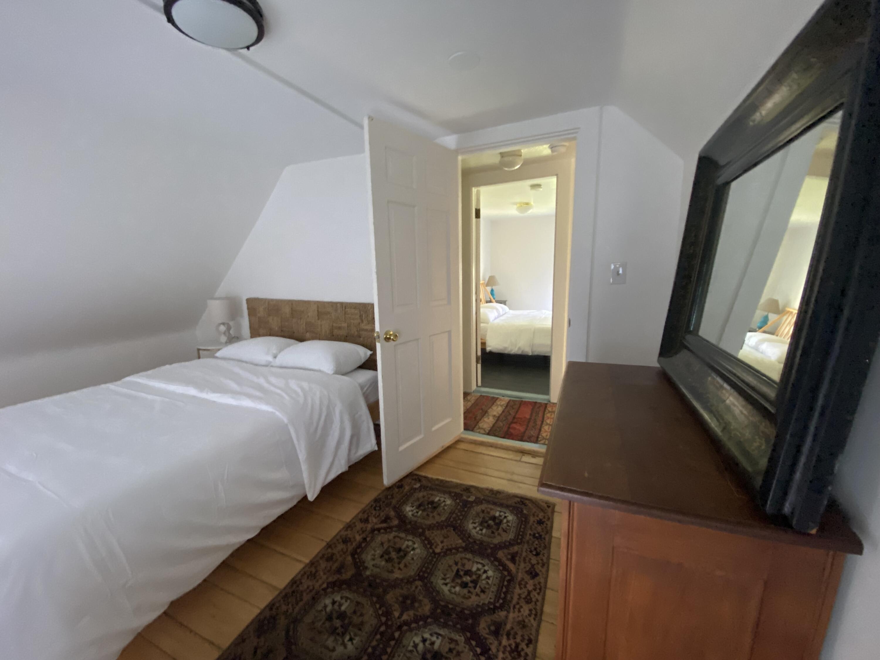 Furnished bedroom with a white bed, sloped ceiling, patterned rug, and doorway to another bedroom.