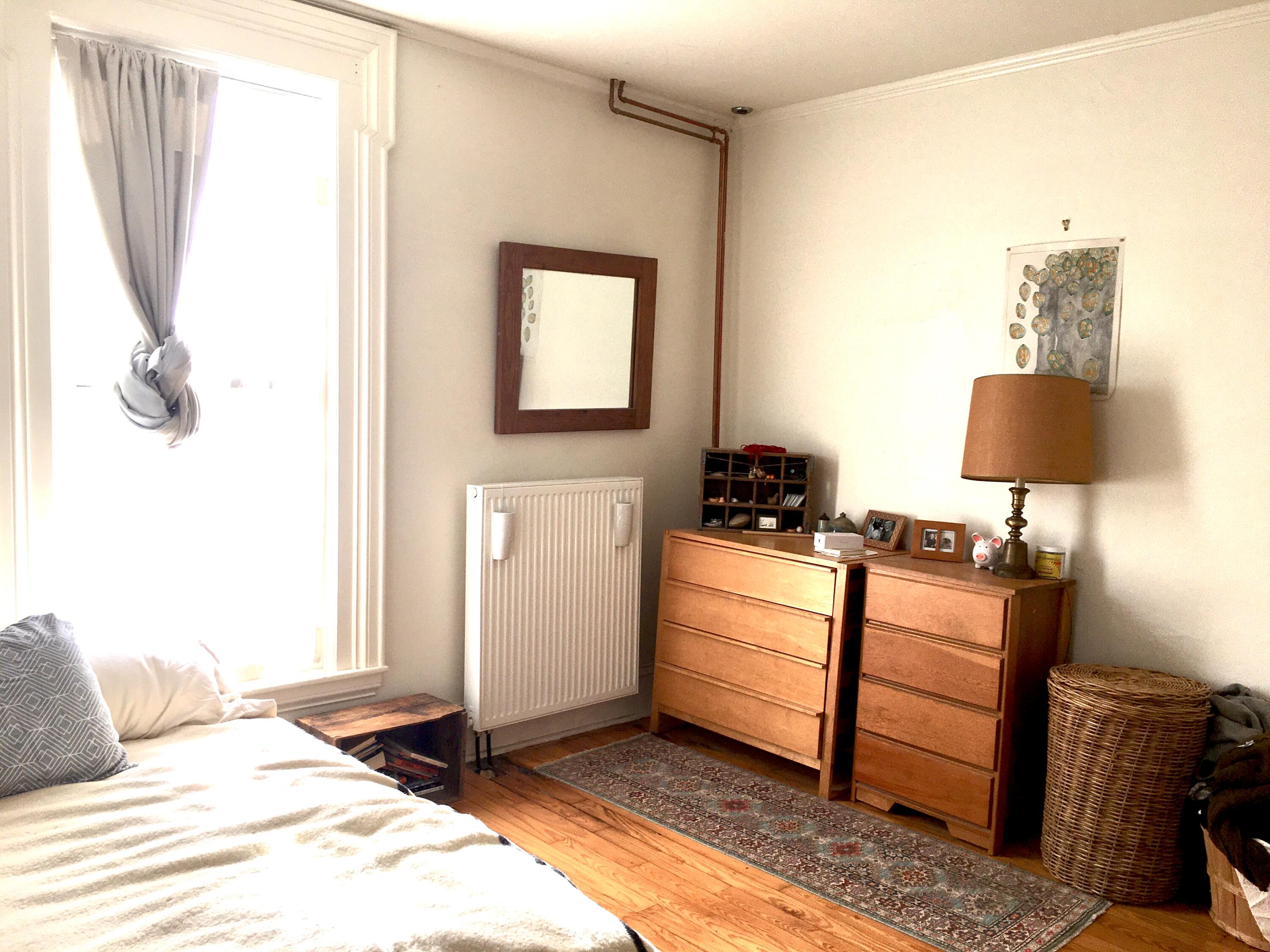 Furnished bedroom with wooden dressers, a radiator, and a bright curtained window.