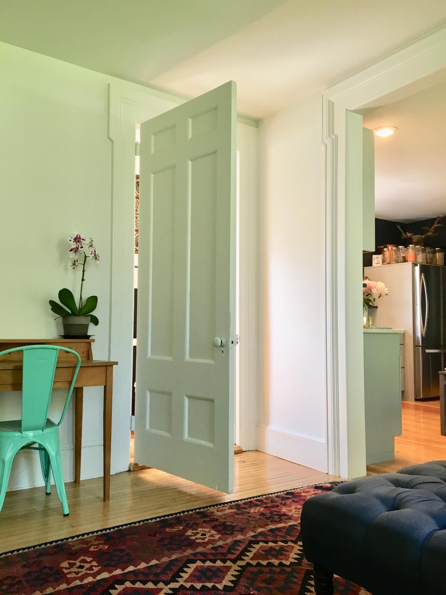 Living room with an open green paneled door, patterned rug, turquoise chair, and kitchen visible beyond.