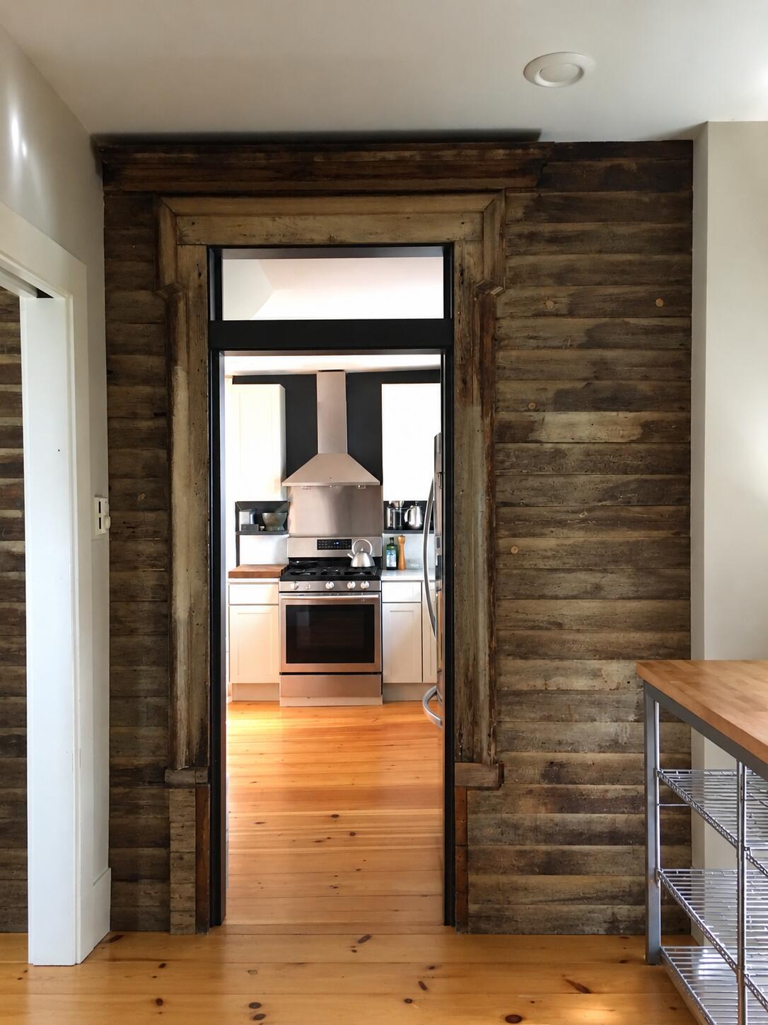 Kitchen beyond a rustic timber doorway, with white cabinets, stainless range, and warm wood floors.
