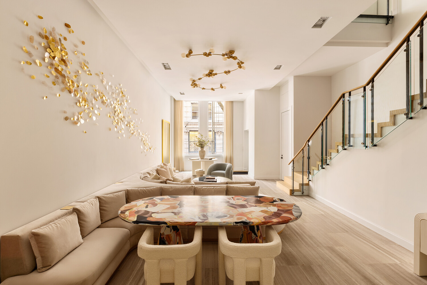 Open-plan living and dining room with a sculptural table, gold wall art, and glass-railed staircase.