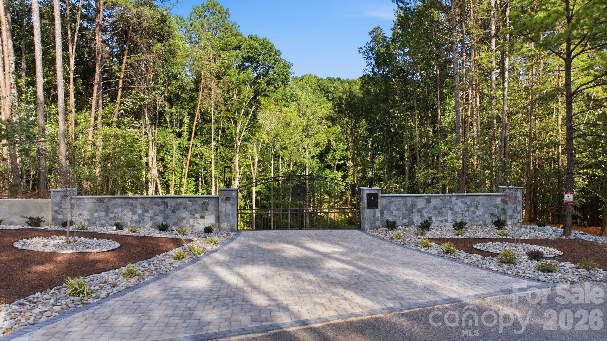 Gated driveway entrance with a black metal gate, stone walls, and dense trees beyond.