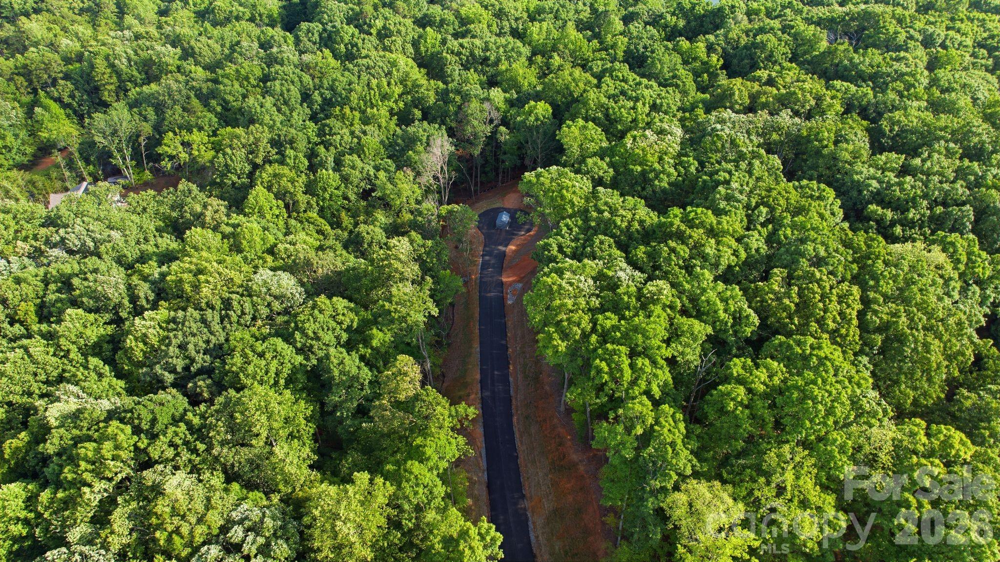 Aerial view of a paved driveway through dense woodland toward a small property site.