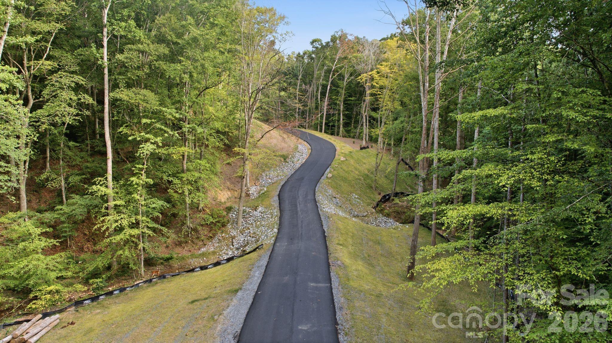 Winding paved road curves through a wooded property with grassy, stone-lined slopes.