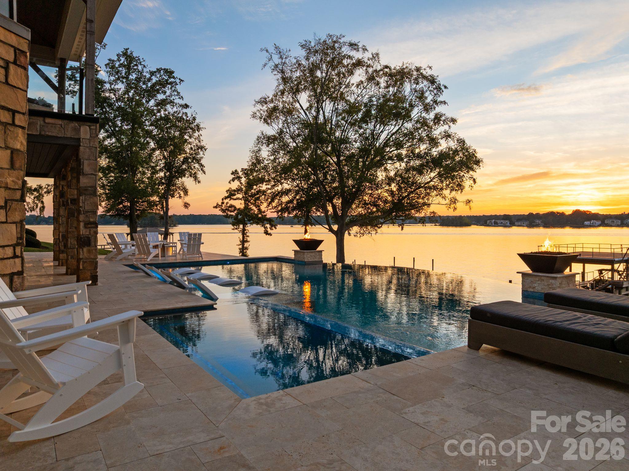 Lakeside pool and stone patio at sunset, with Adirondack chairs and fire features beside the water.