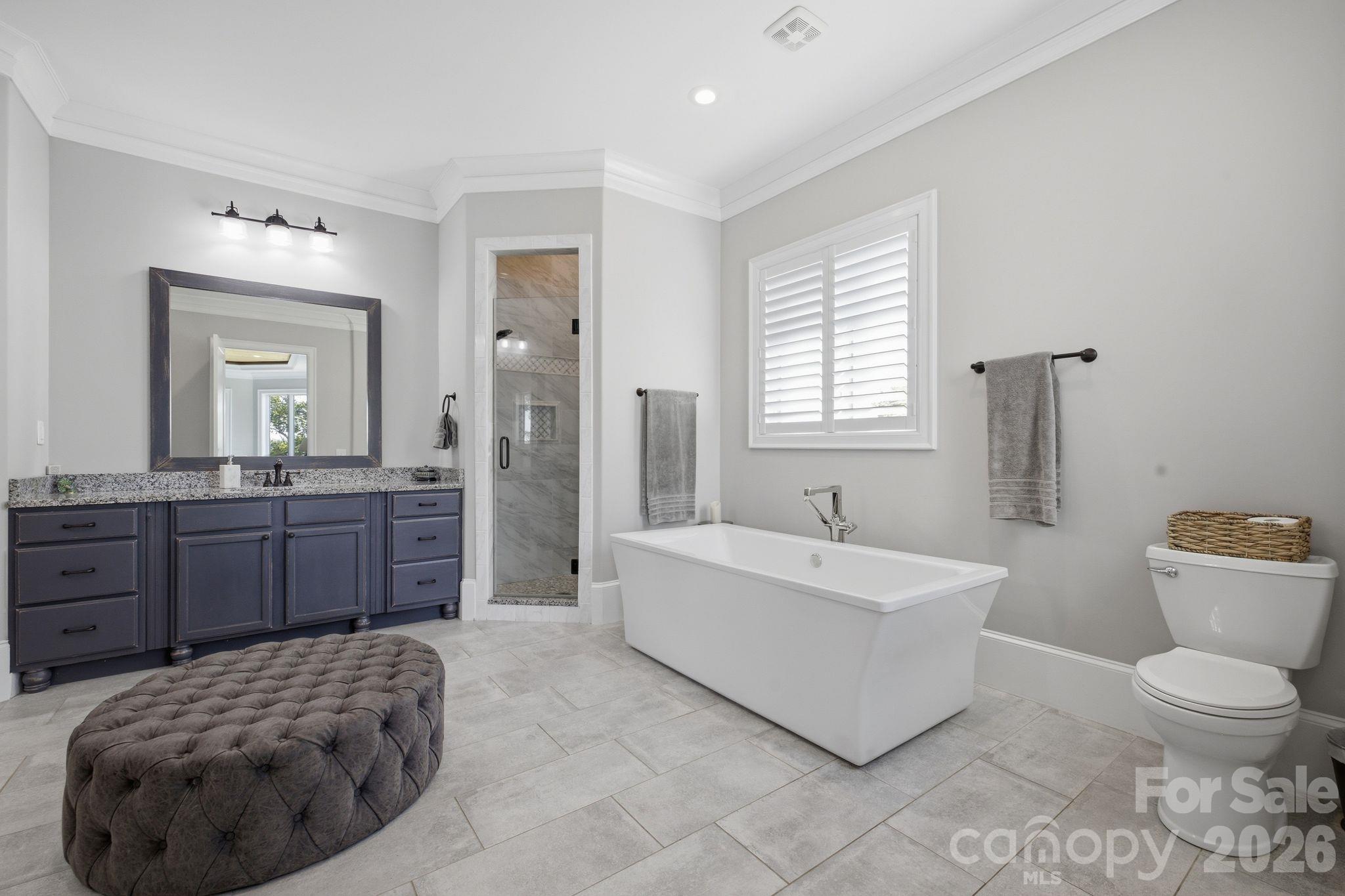Bathroom with freestanding tub, glass shower, dark double vanity, and tufted ottoman.