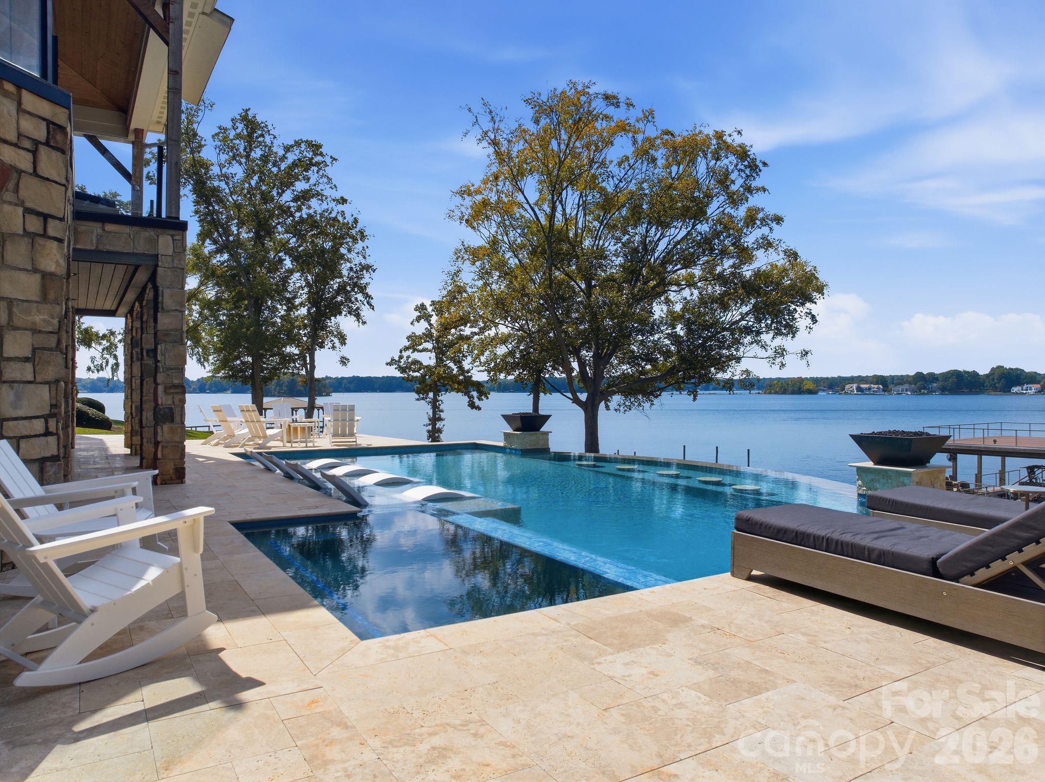 Lakeside pool and stone patio with lounge chairs, mature trees, and a view across the water.