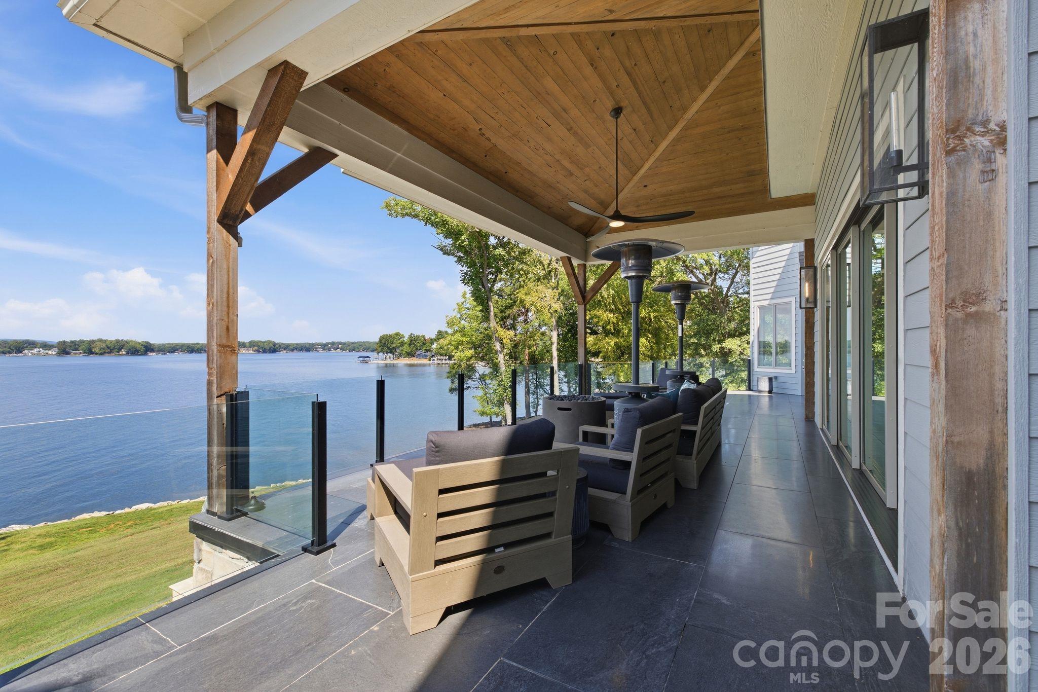 Covered lakeside patio with lounge seating, glass railing, and a view over the water.