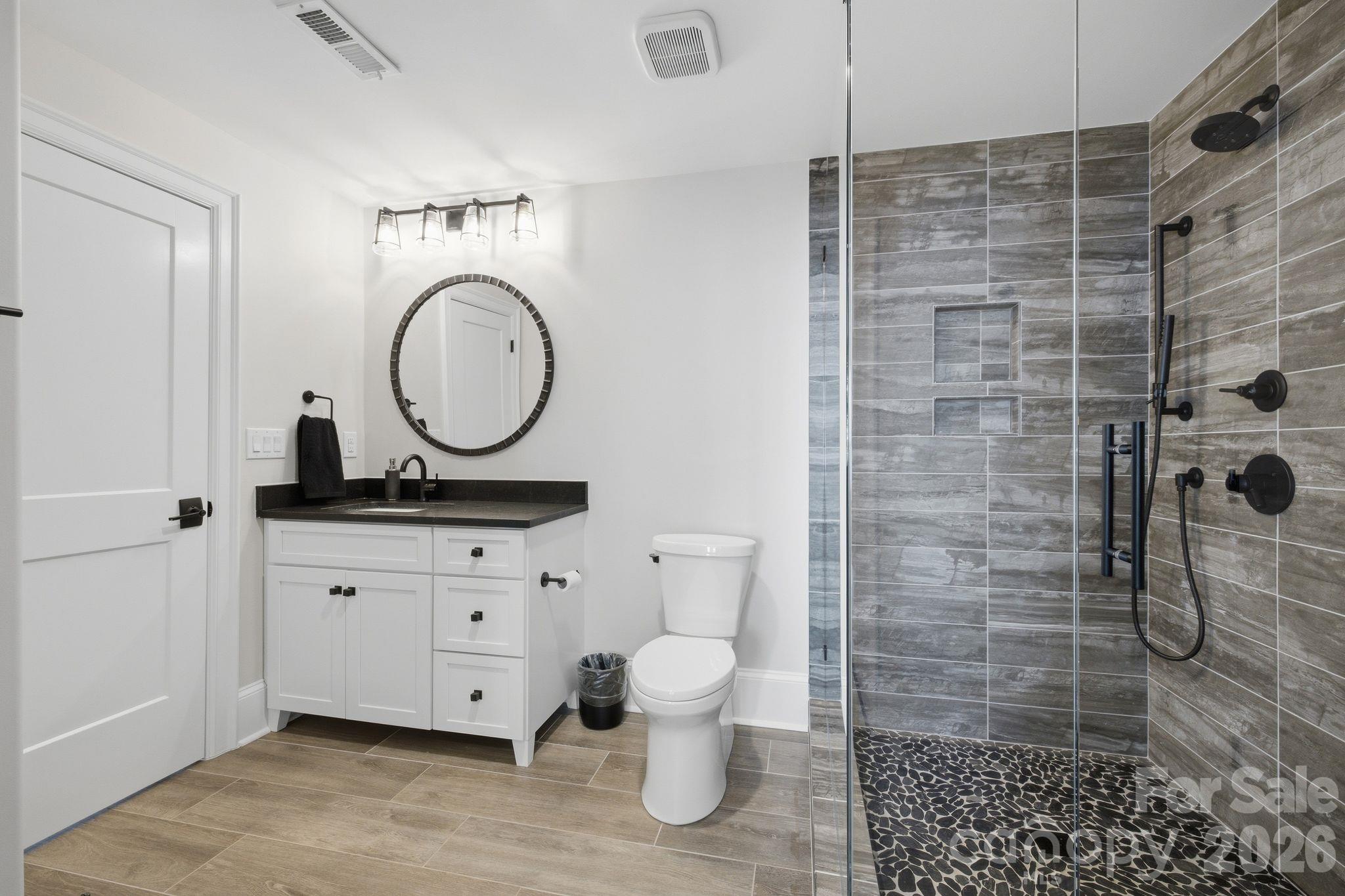 Bathroom with a white vanity and round mirror beside a glass shower with gray tile and a pebble floor.