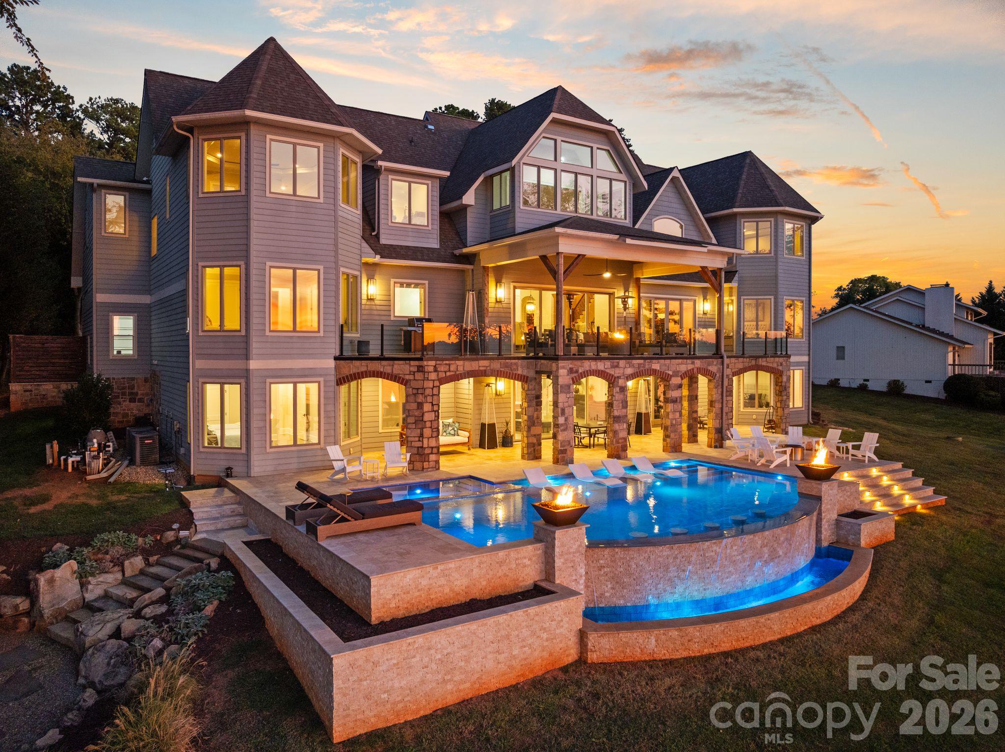 Large gray-shingled home with a stone terrace and illuminated swimming pool at sunset.