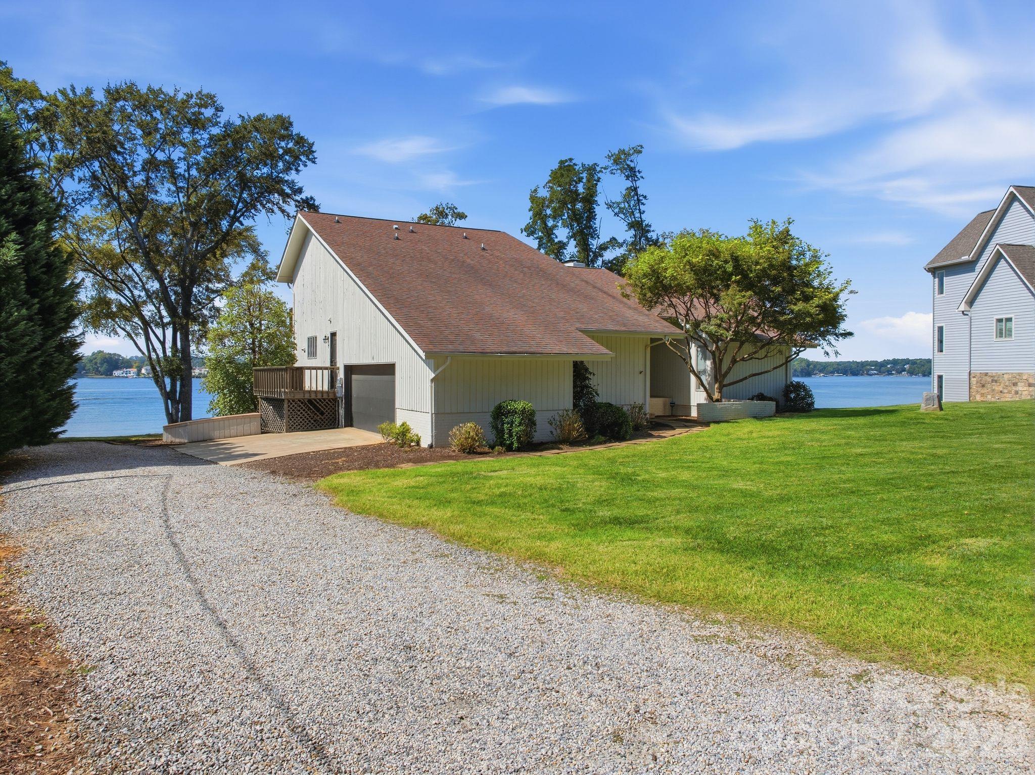 Lakeside home with a gravel driveway, green lawn, small deck, and lake visible behind it.