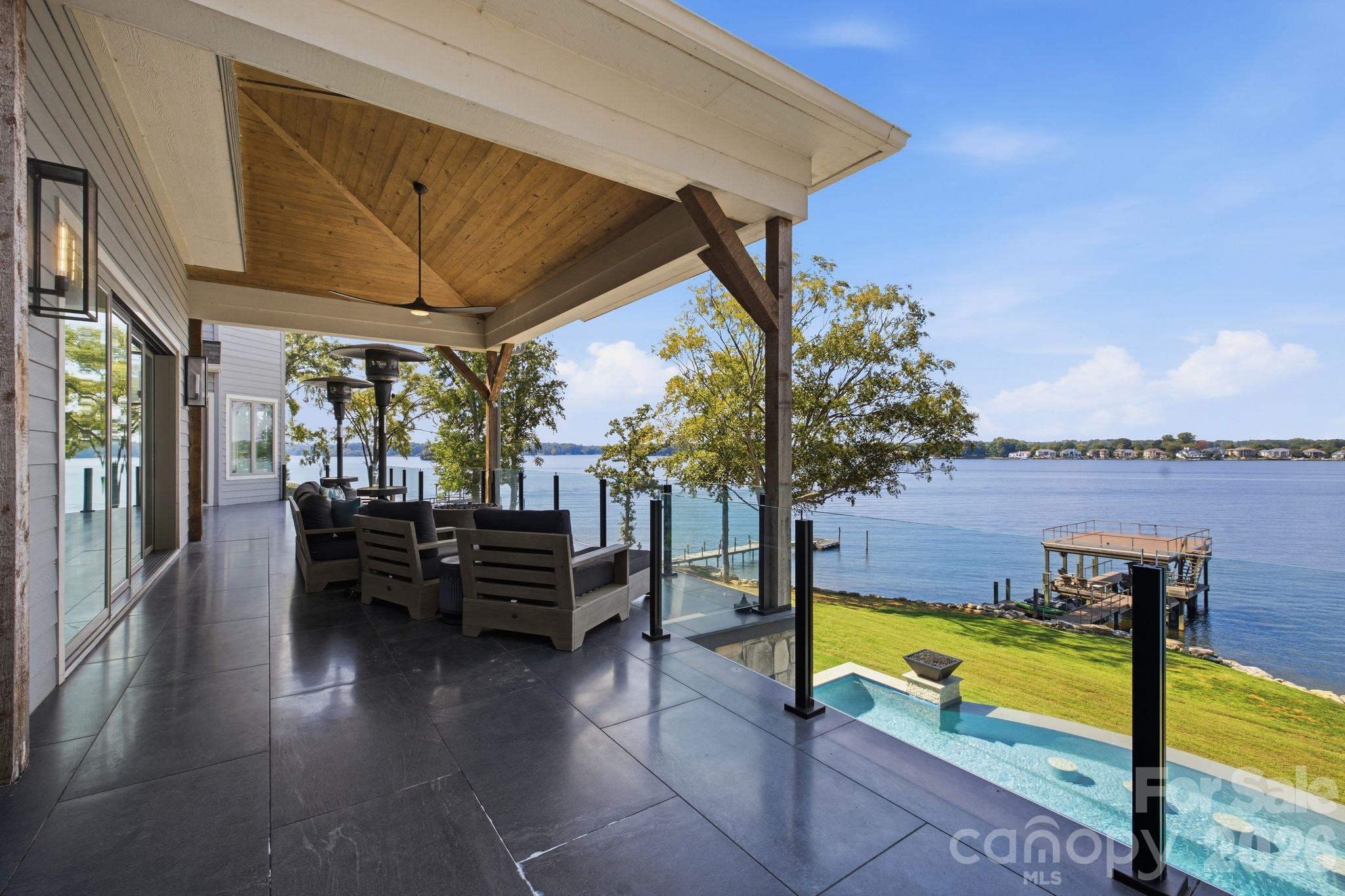 Covered lakeside patio with outdoor seating, glass railing, pool, and dock overlooking the water.