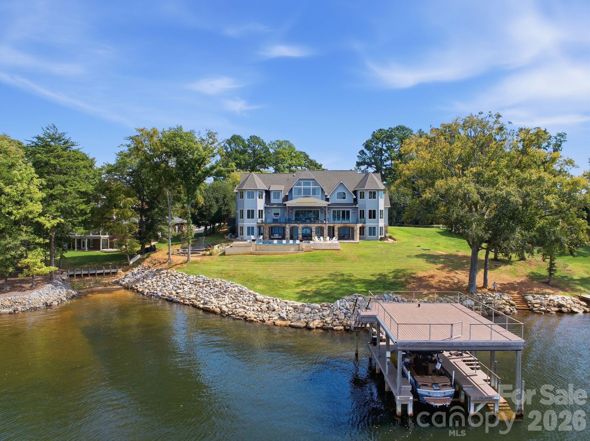 Lakeside home with a tree-lined lawn, rocky shore, and dock with a boat.