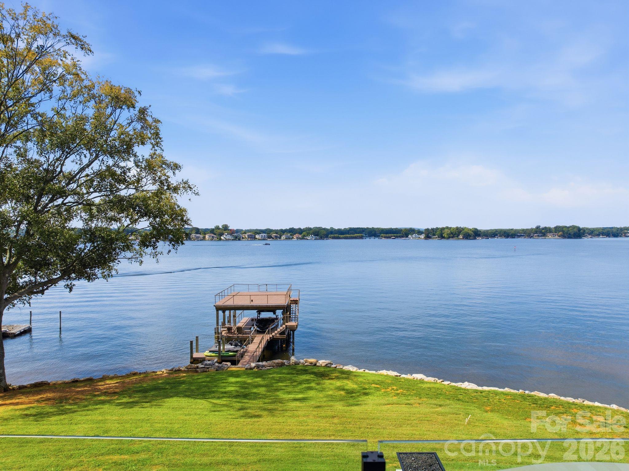 Lake view with a covered boat dock, grassy shoreline, and homes across the water.