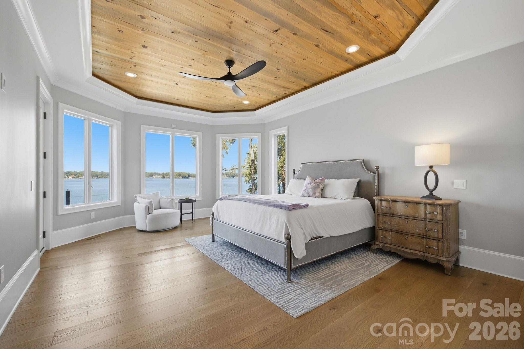 Staged bedroom with a wood-plank tray ceiling and large windows overlooking the water.