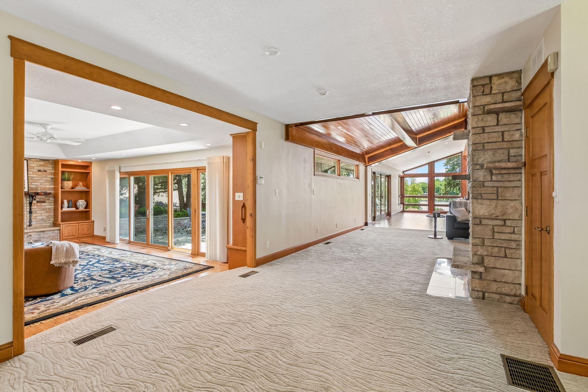 Spacious carpeted living room with a stone fireplace, vaulted wood ceiling, and large windows.