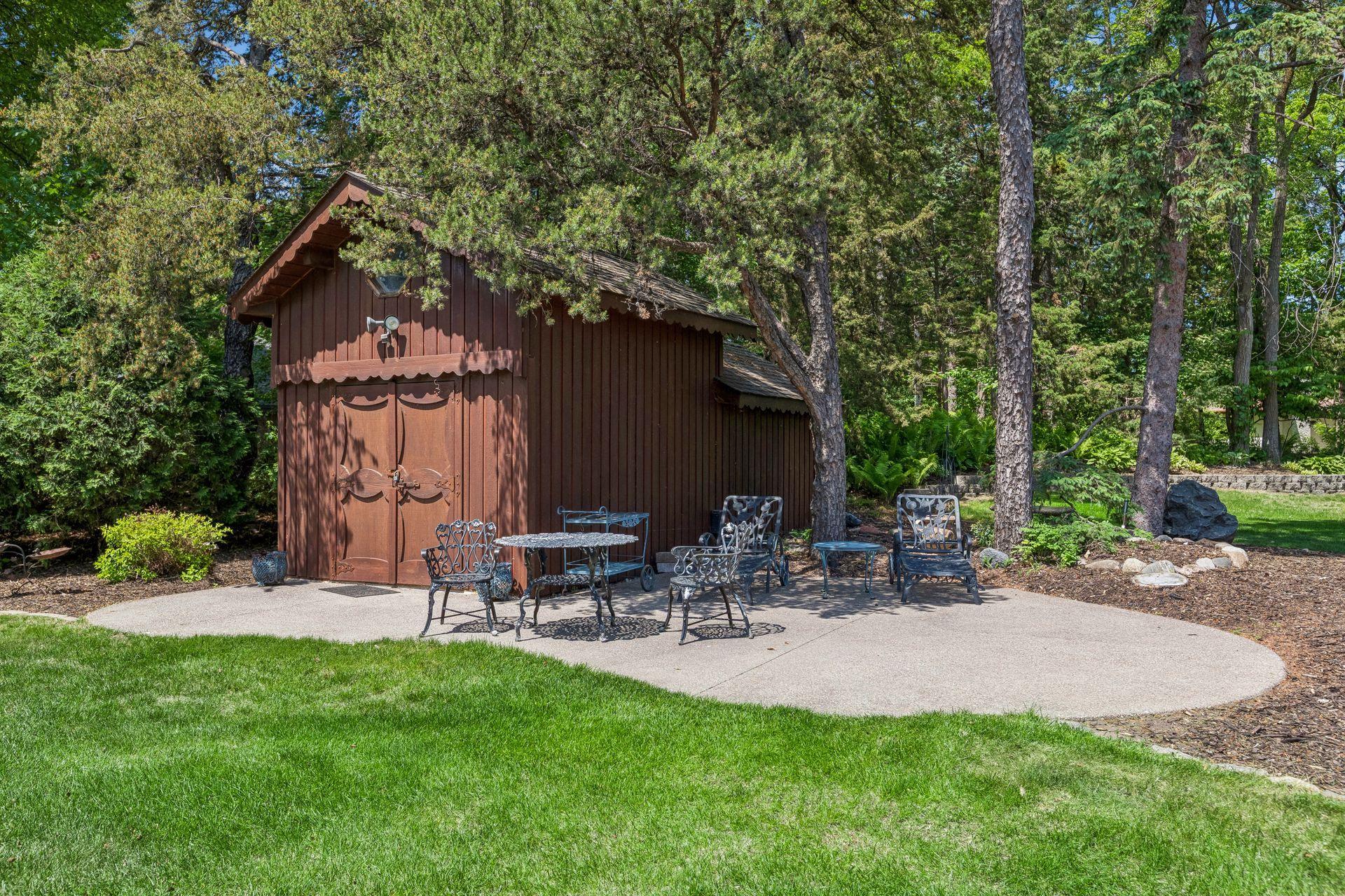 Brown wooden outbuilding and metal patio seating beneath mature trees beside a curved concrete patio.