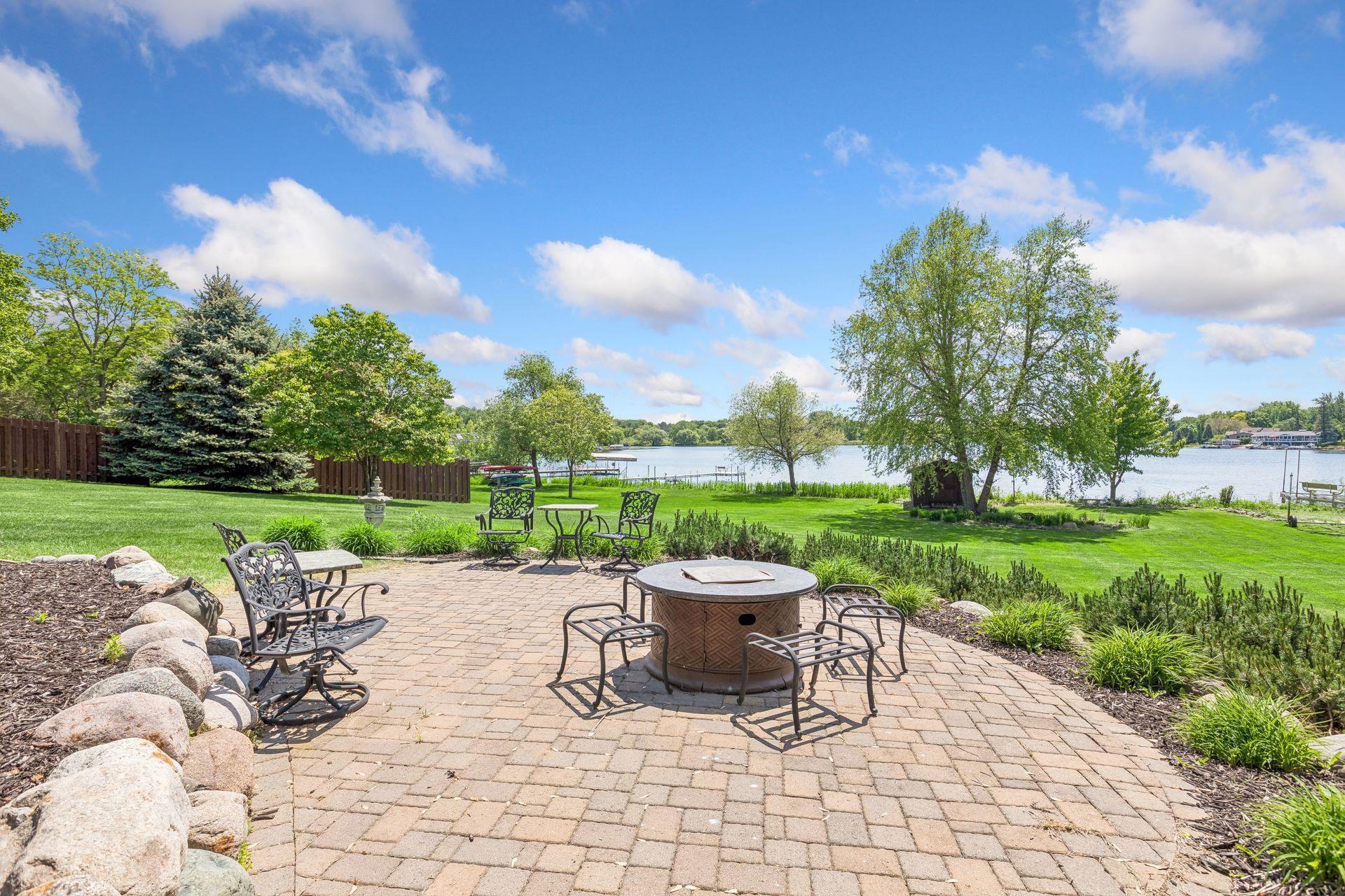 Waterfront patio with a round fire pit and metal seating overlooking a green lawn and lake.